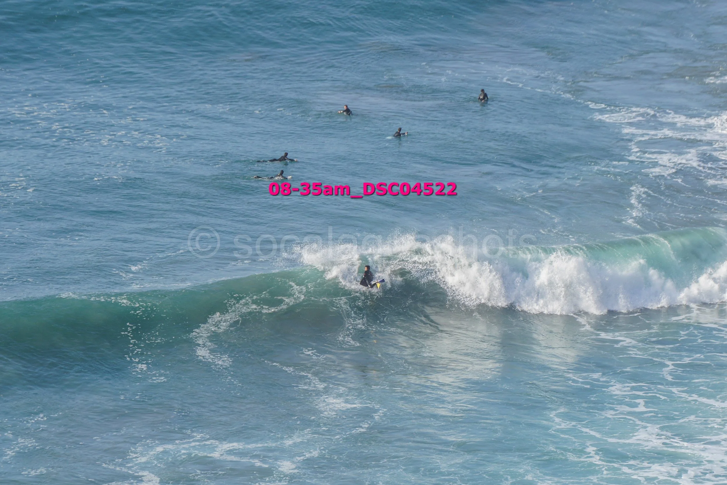 A group of surfers in the ocean, with one surfer riding a wave and several others waiting or paddling further out in the water.