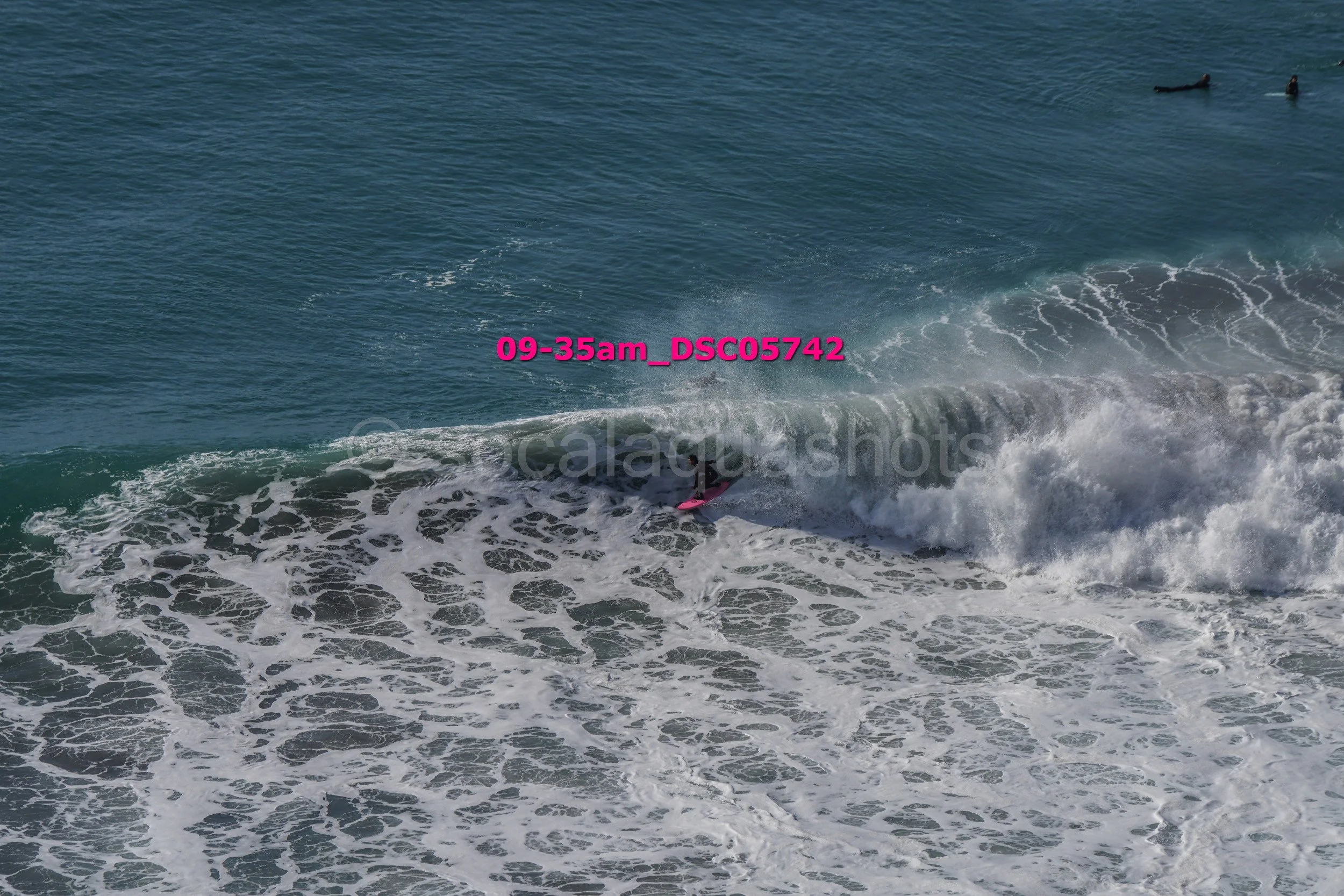 A person surfing on a pink surfboard on a breaking wave in the ocean, with two other surfers visible in the water in the distance.