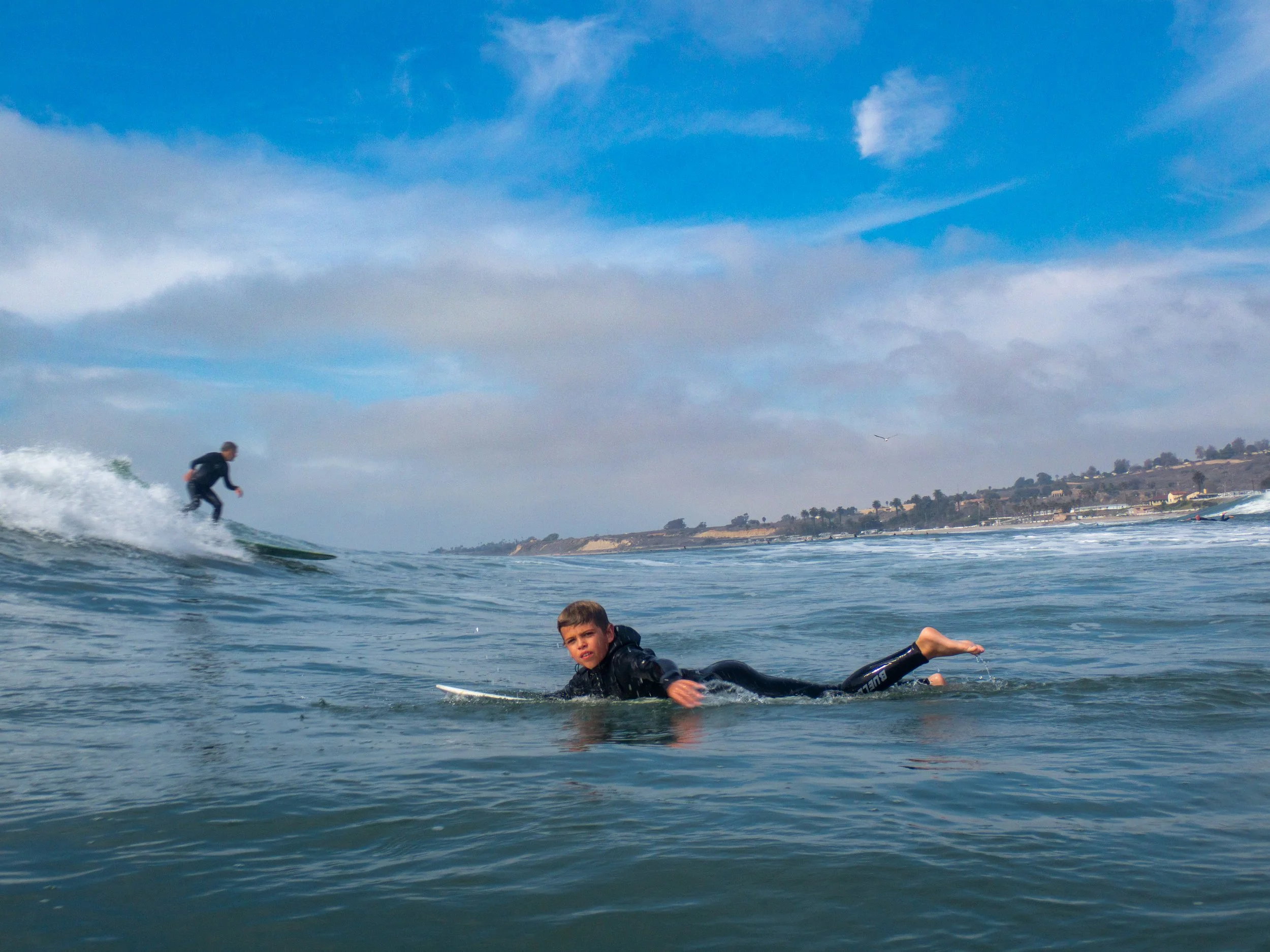 A boy in a wetsuit lying on a surfboard in the water, with another boy riding a wave in the background at the beach under a partly cloudy sky.