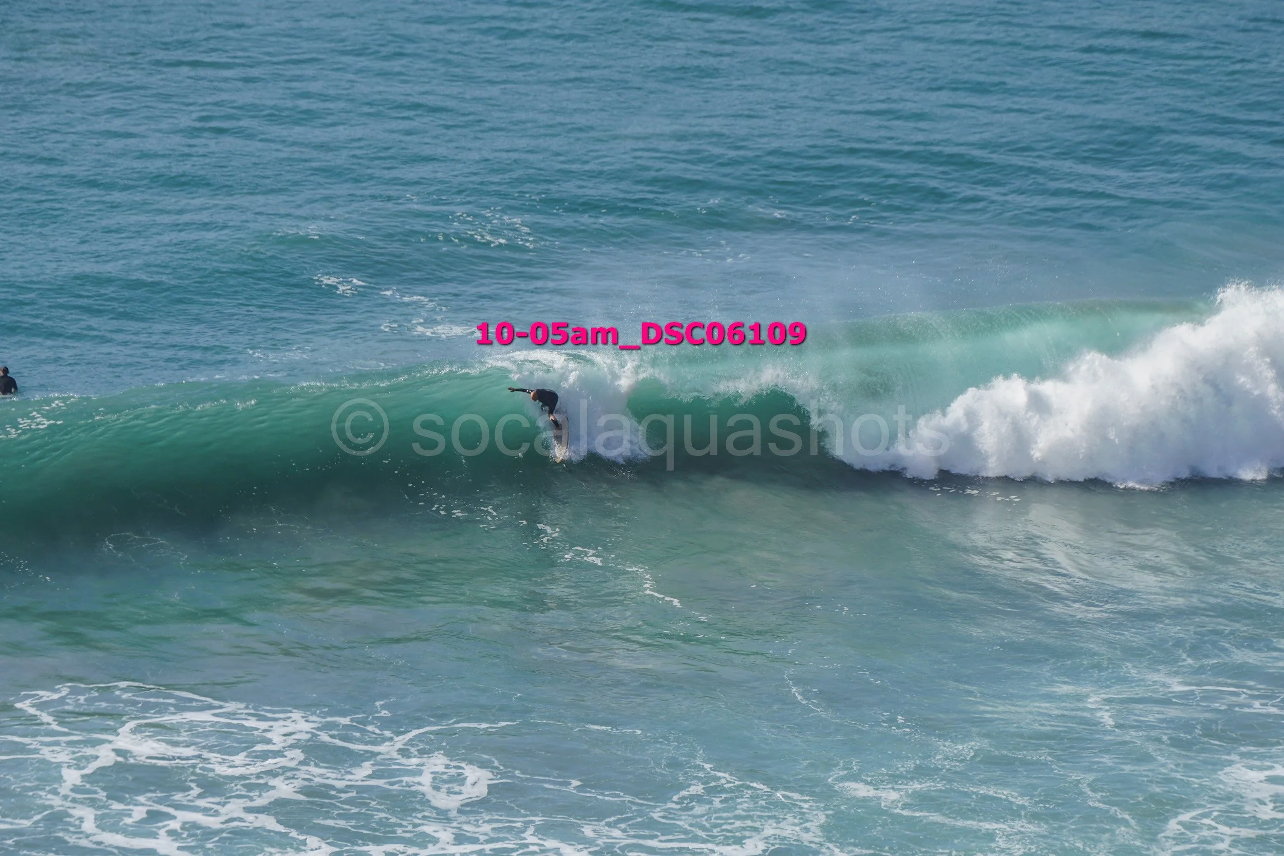 A surfer riding inside the wave with the ocean in the background, another person is visible in the water to the left.