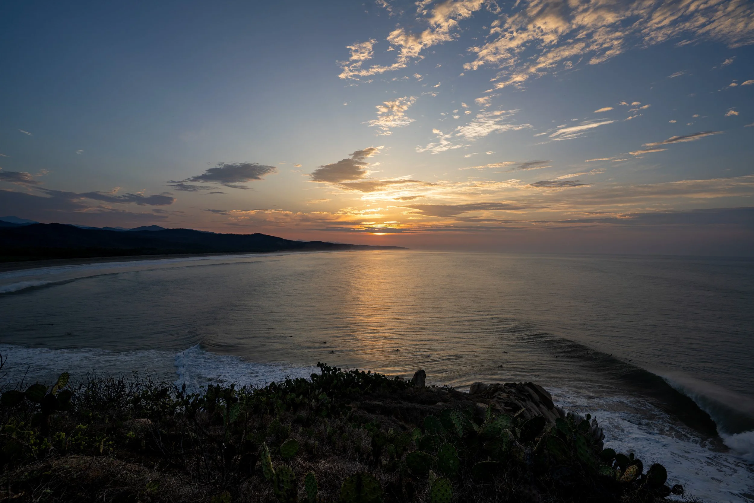 Sunset over the ocean with a partly cloudy sky, mountains in the distance, and Coastal vegetation and rocks in the foreground.