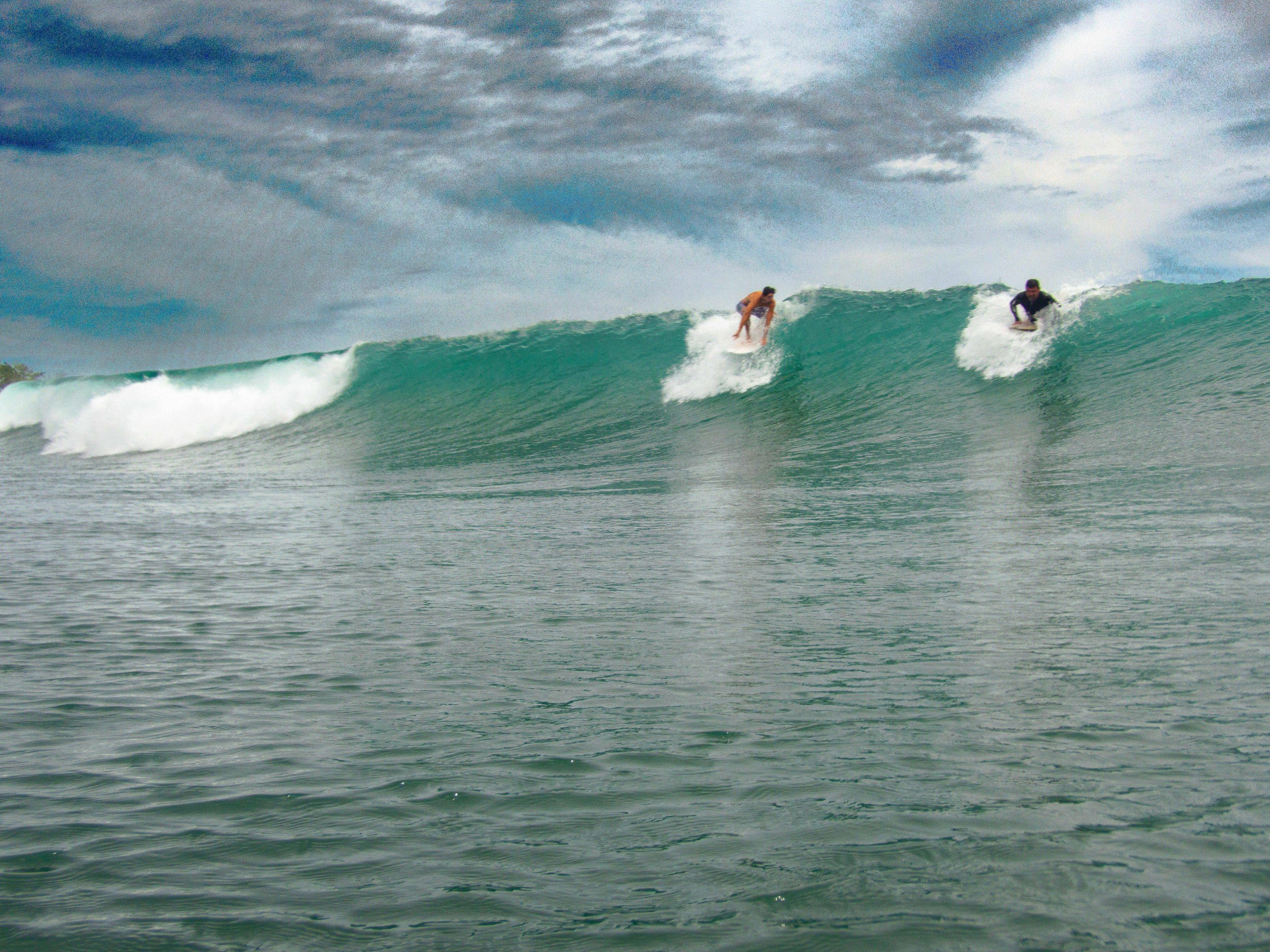 two surfers riding a large ocean wave under a cloudy sky