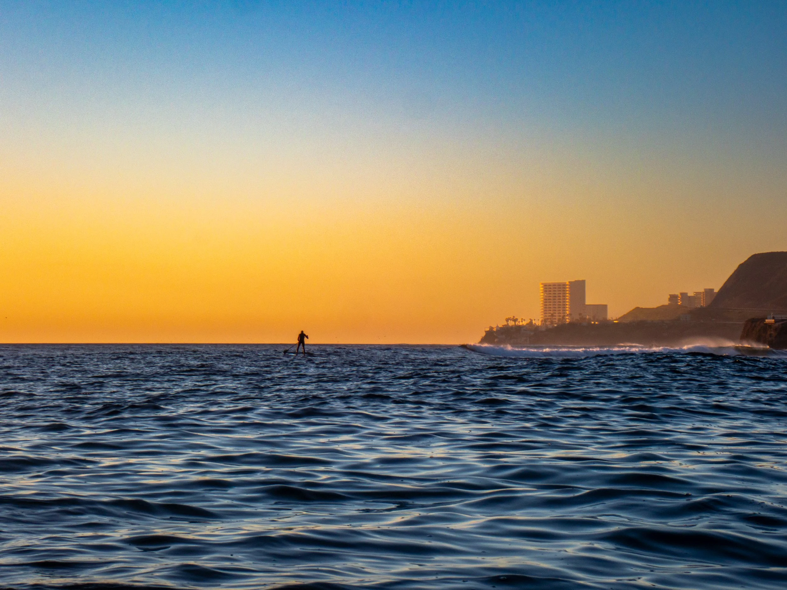 A person paddleboarding on the ocean during sunset with buildings and hills in the distance.