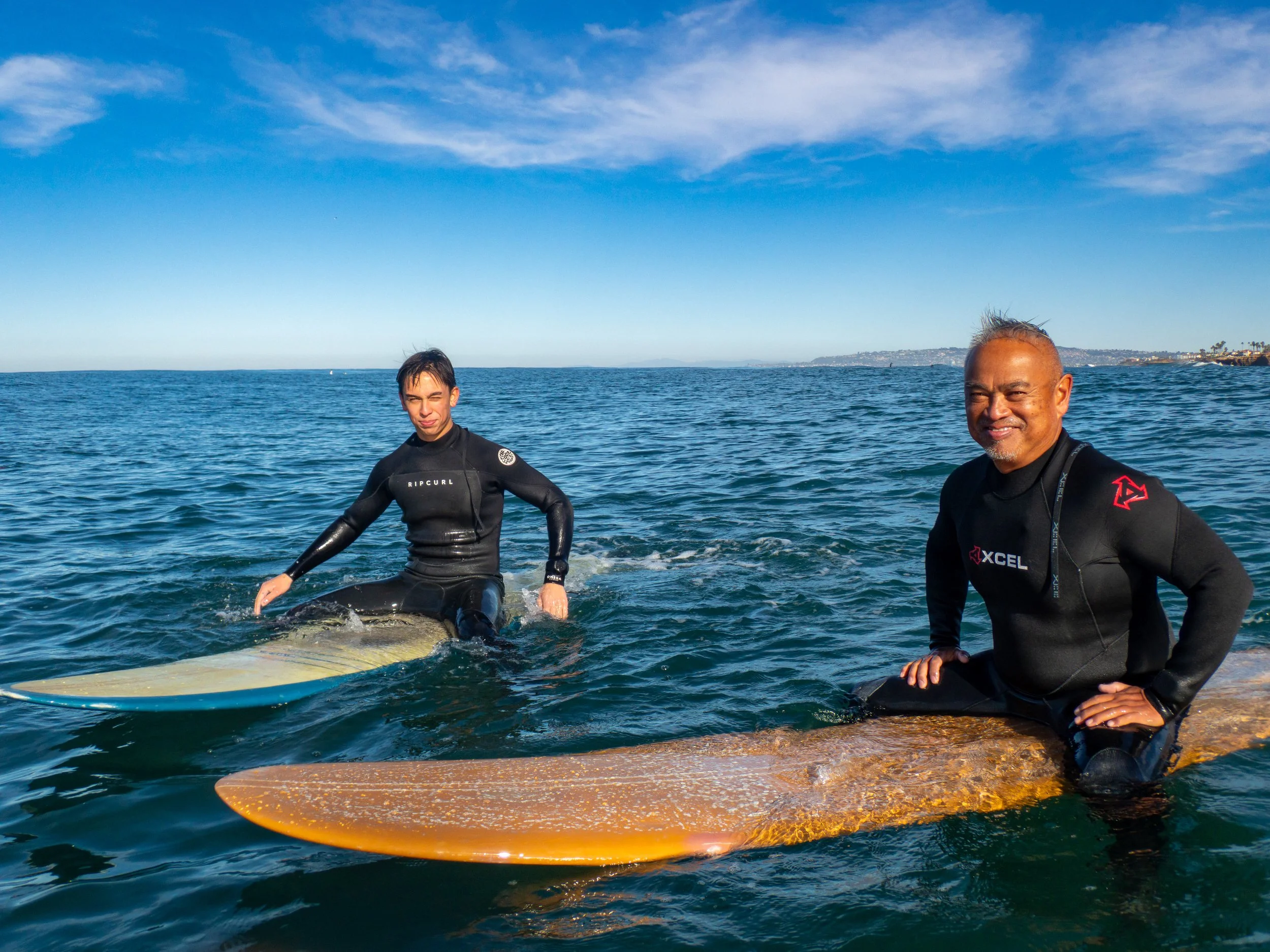 Two surfers, one younger man and one older man, sitting on surfboards in the ocean with a clear blue sky and distant shoreline in the background.