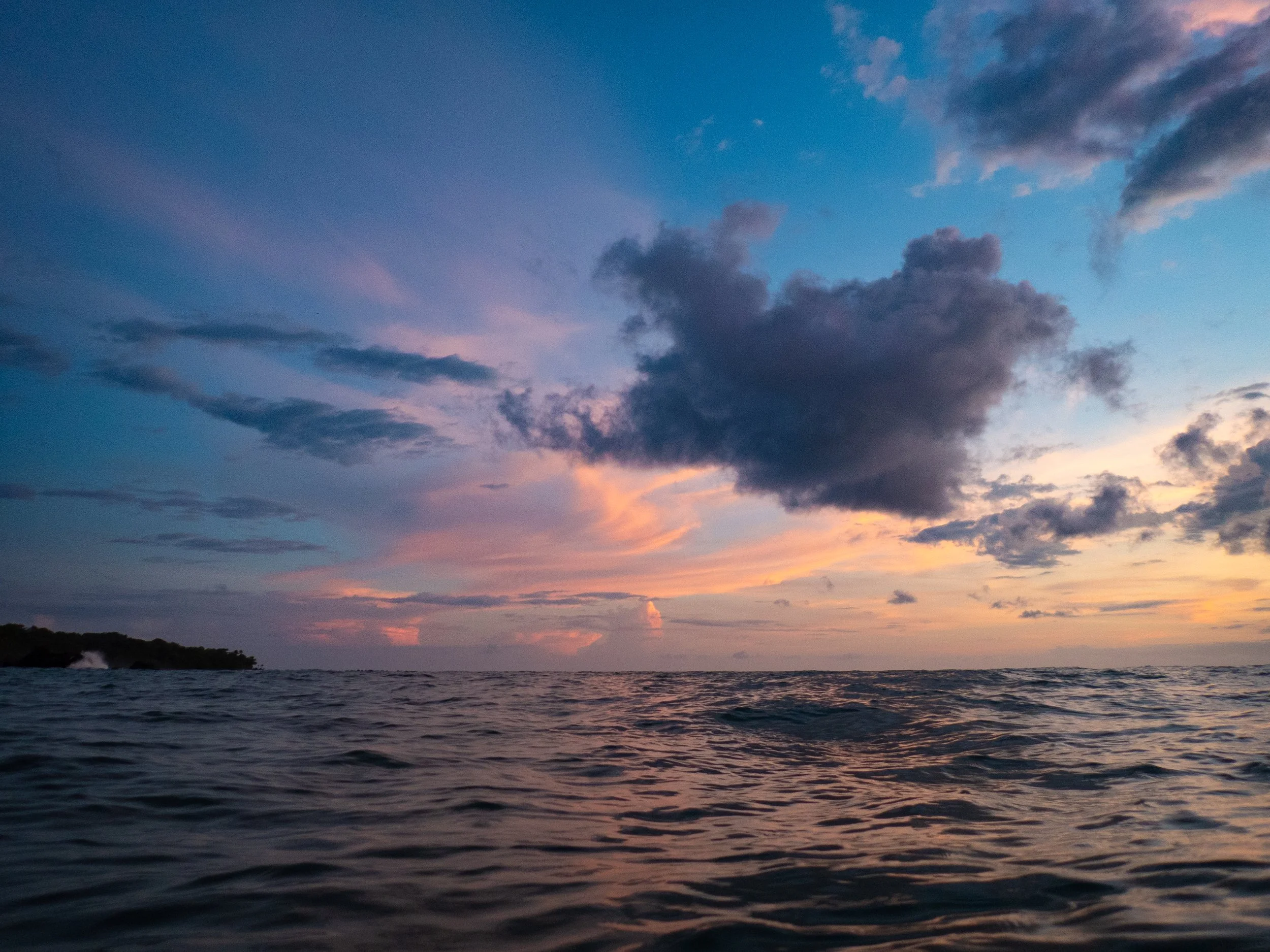 A view of the ocean at sunset with dark clouds in the sky and a small landmass in the distance.