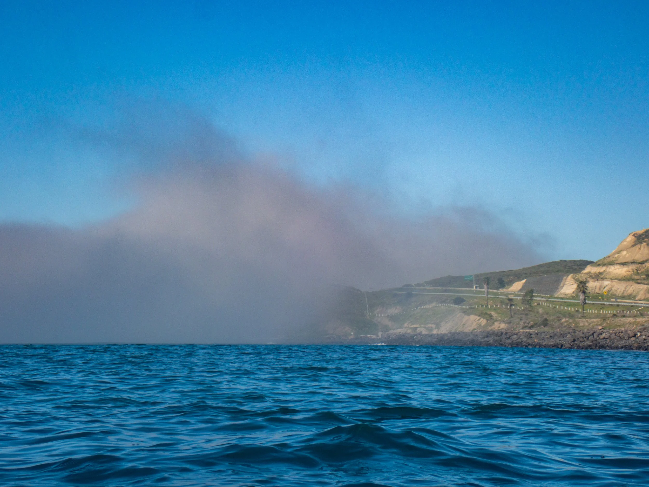 Ocean water with distant foggy coastline, cliffs, and a road on the hillside under a blue sky.