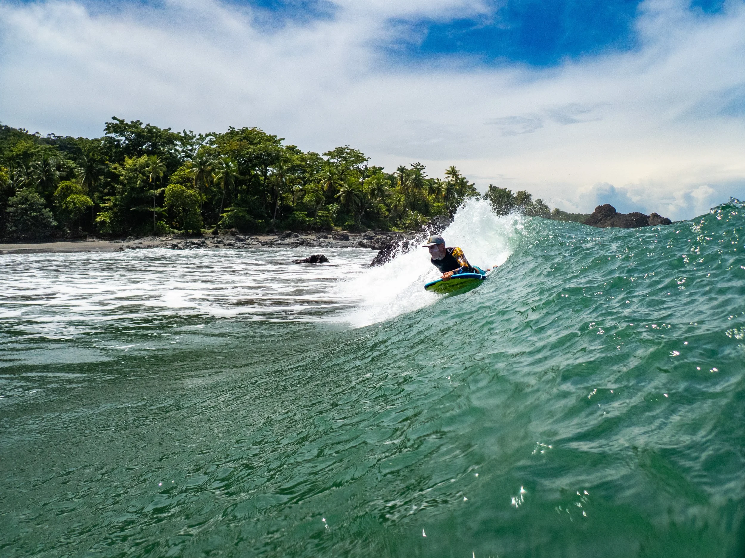 A person surfing on a turquoise wave near a tropical shoreline with green trees under a partly cloudy sky.