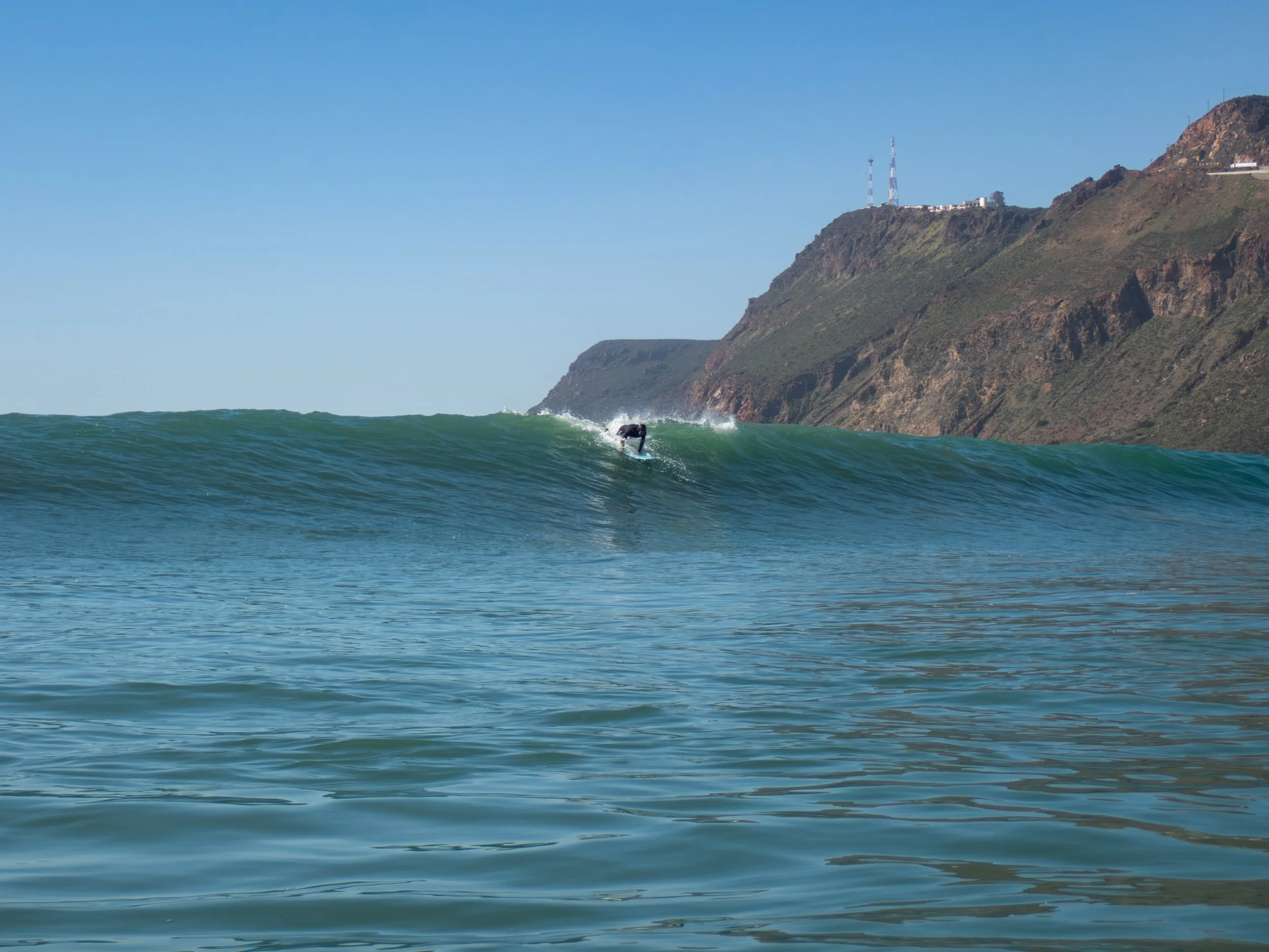 A surfer riding a wave near a coastline with cliffs and communication towers visible in the background on a clear day.