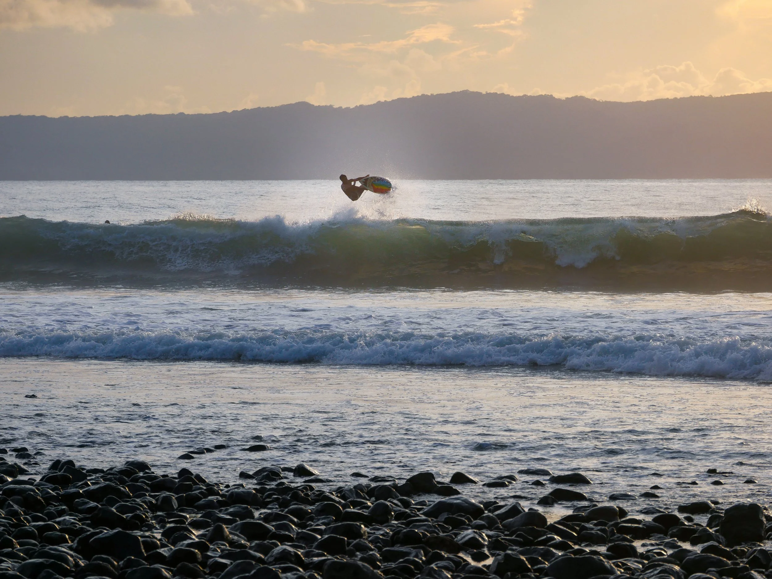 Surfer catching a wave at sunset with rocky beach in foreground and hilly coastline in background