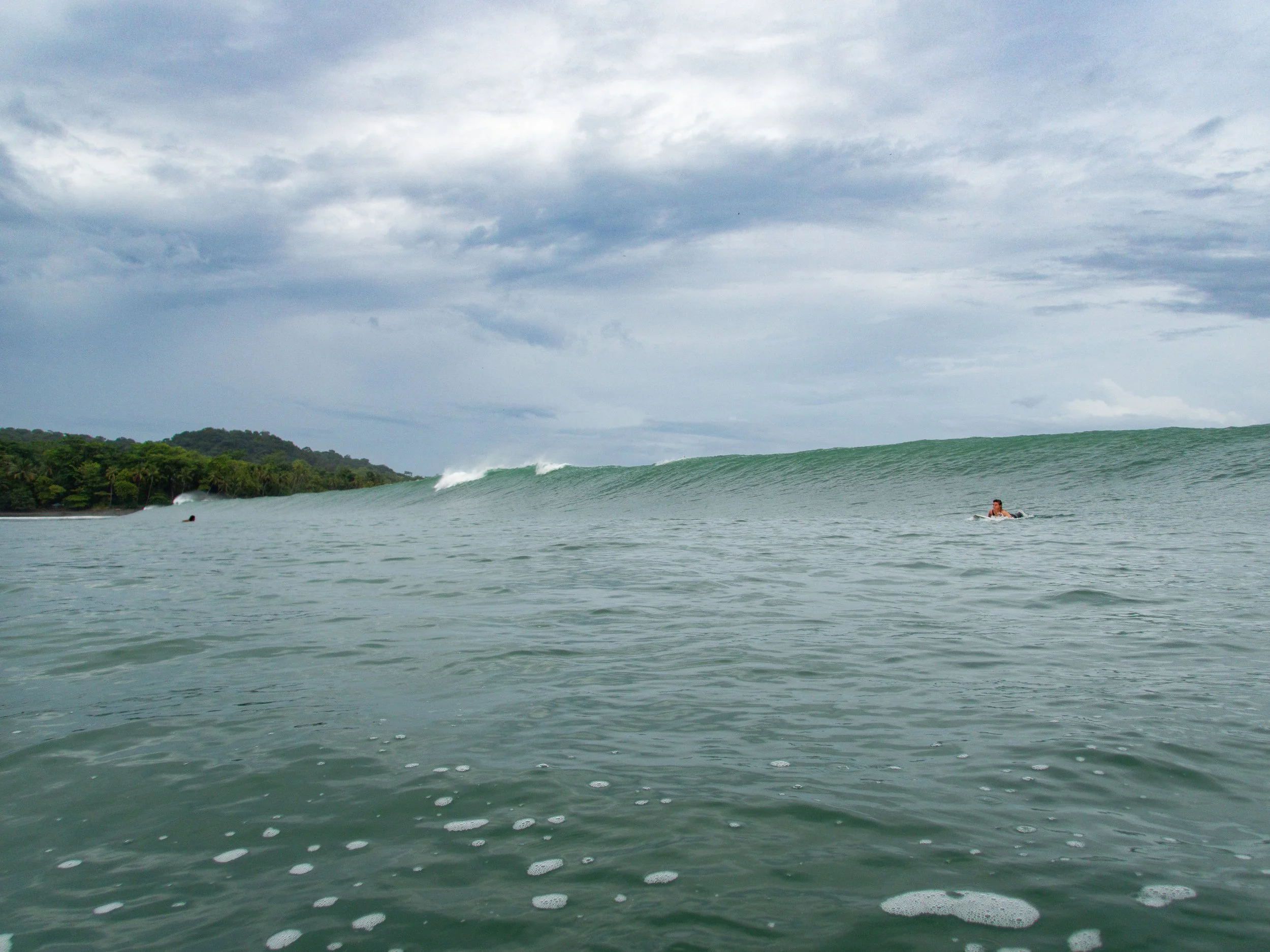 Surfer paddling towards large ocean wave near tropical coastline under cloudy sky.