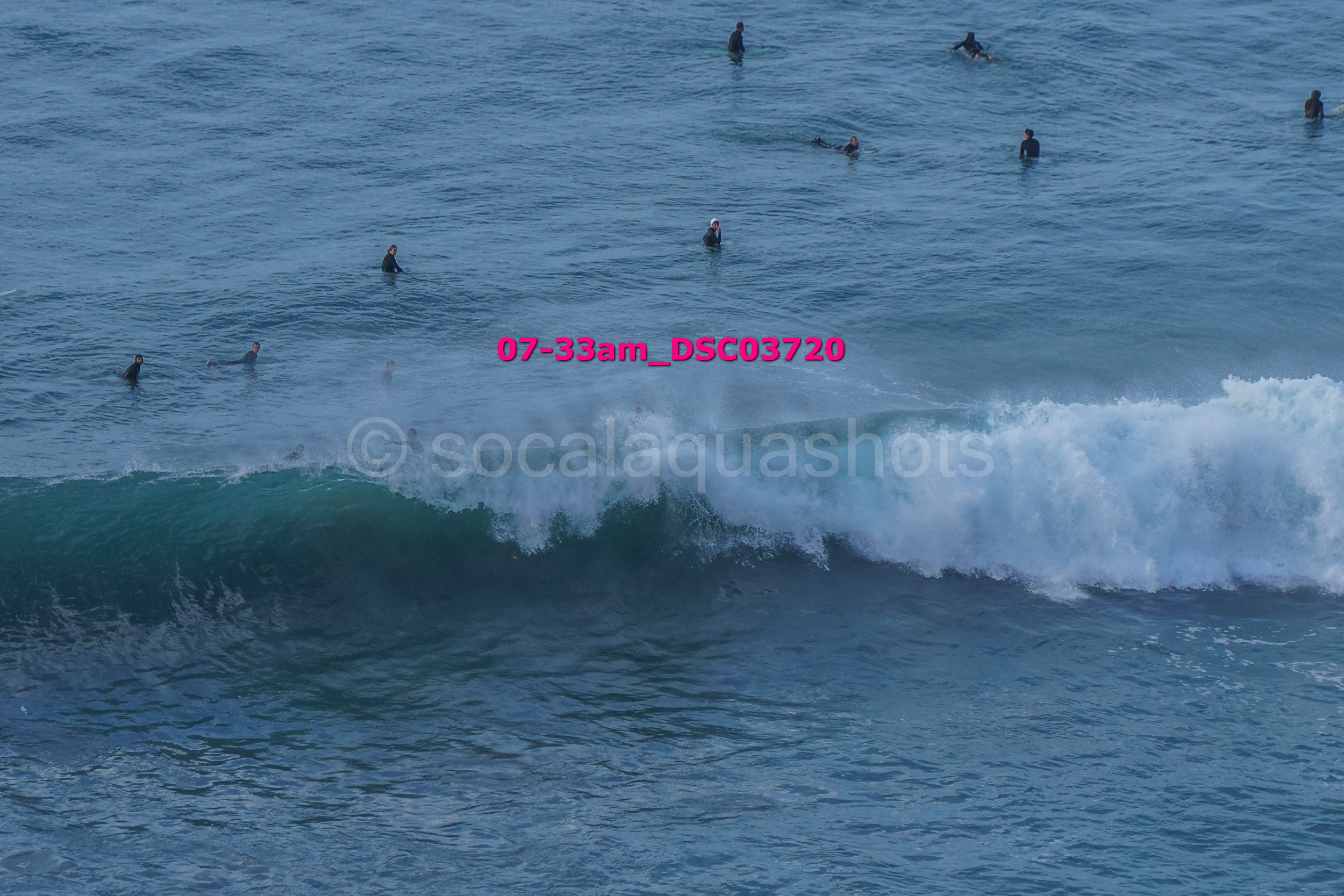 People swimming and surfing in the ocean with a wave in the foreground, some in black wetsuits.