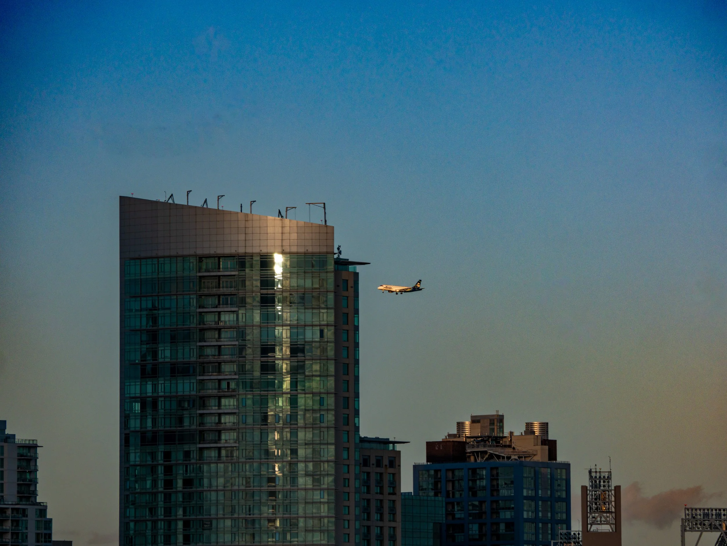 City skyline with tall modern glass building and an airplane flying nearby during sunset or sunrise.