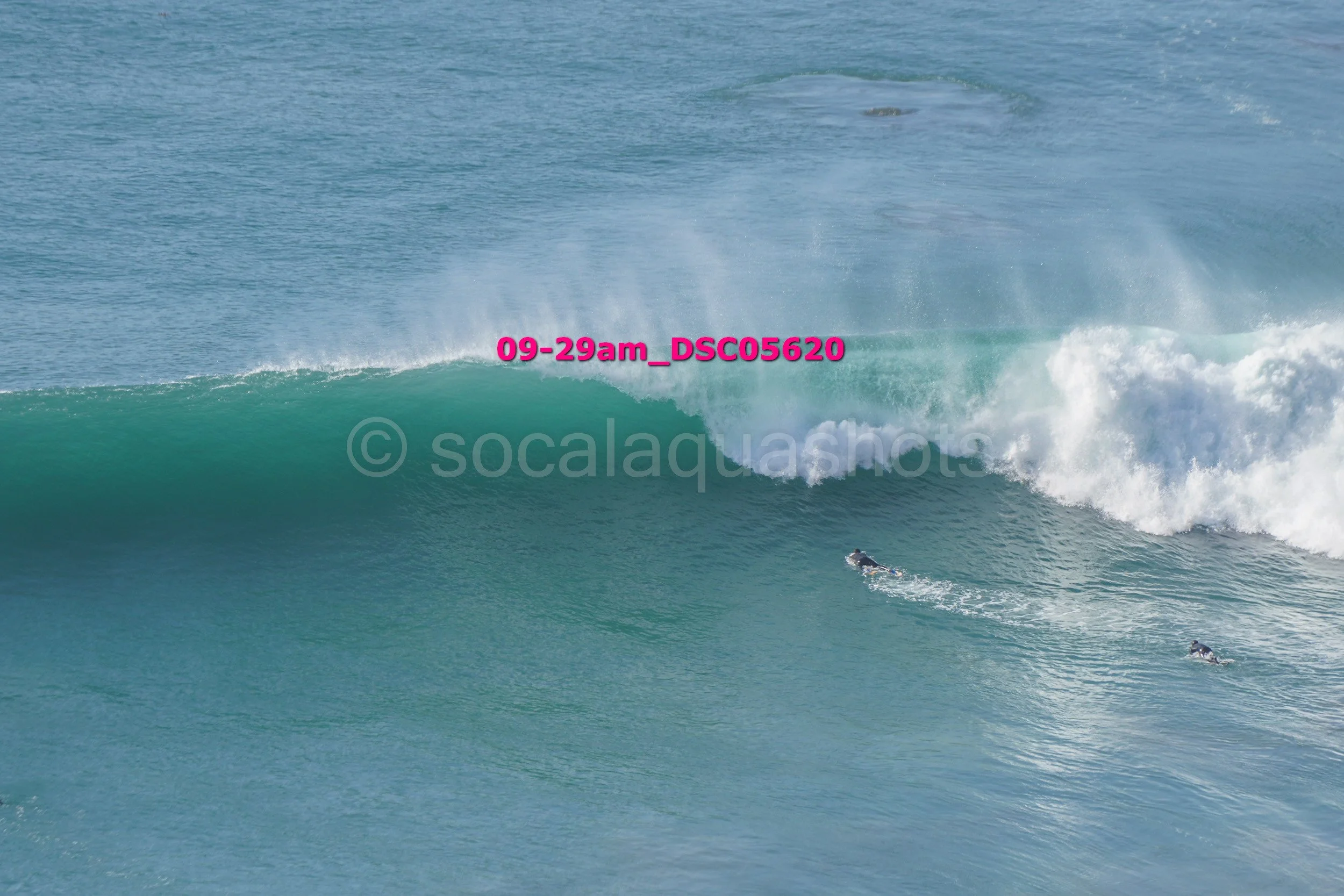 Two surfers riding a large ocean wave with white foam, seen from the side, with a distant water surface in the background.