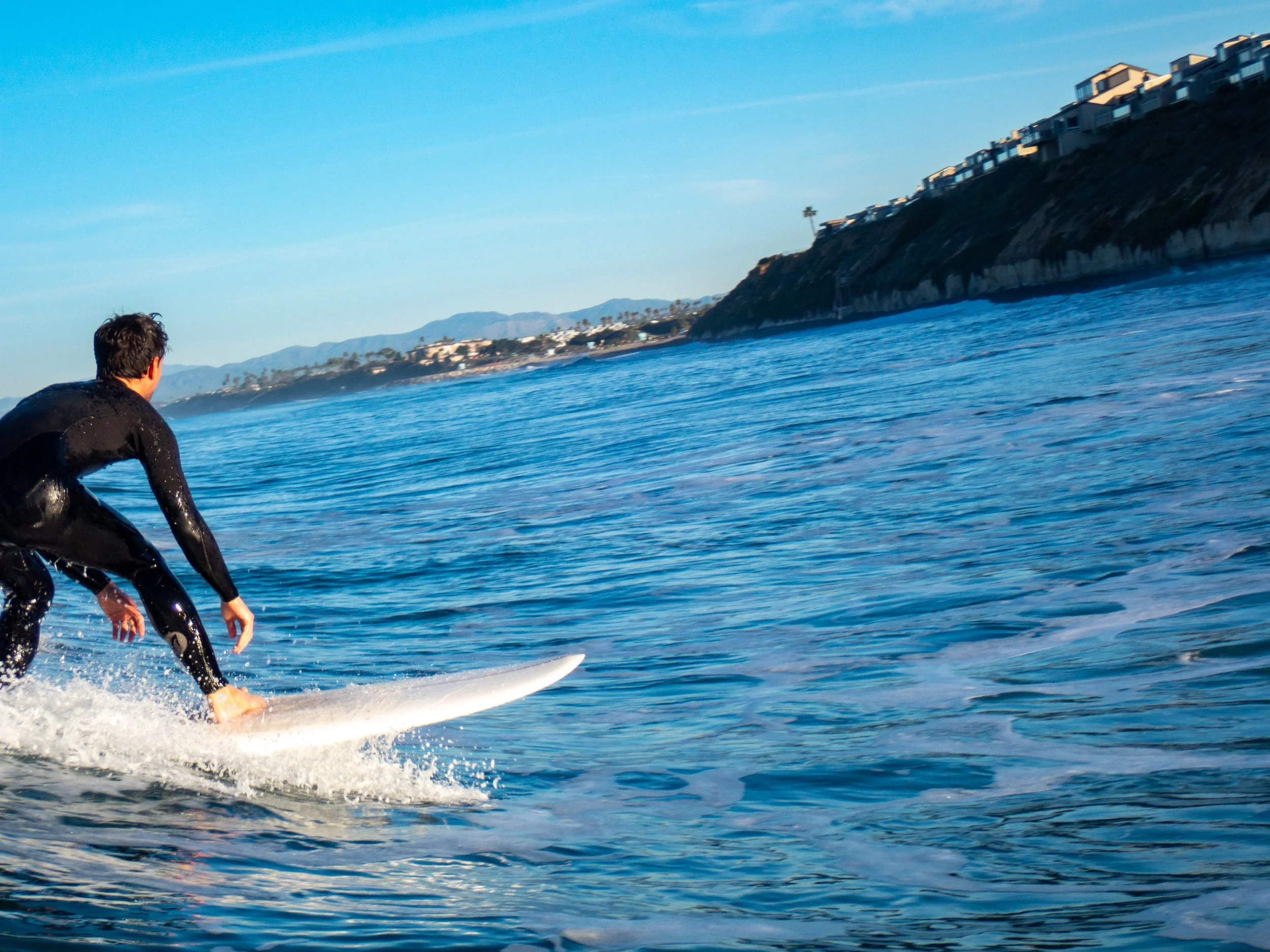 A man surfing in the ocean with a coastal hillside and houses in the background.