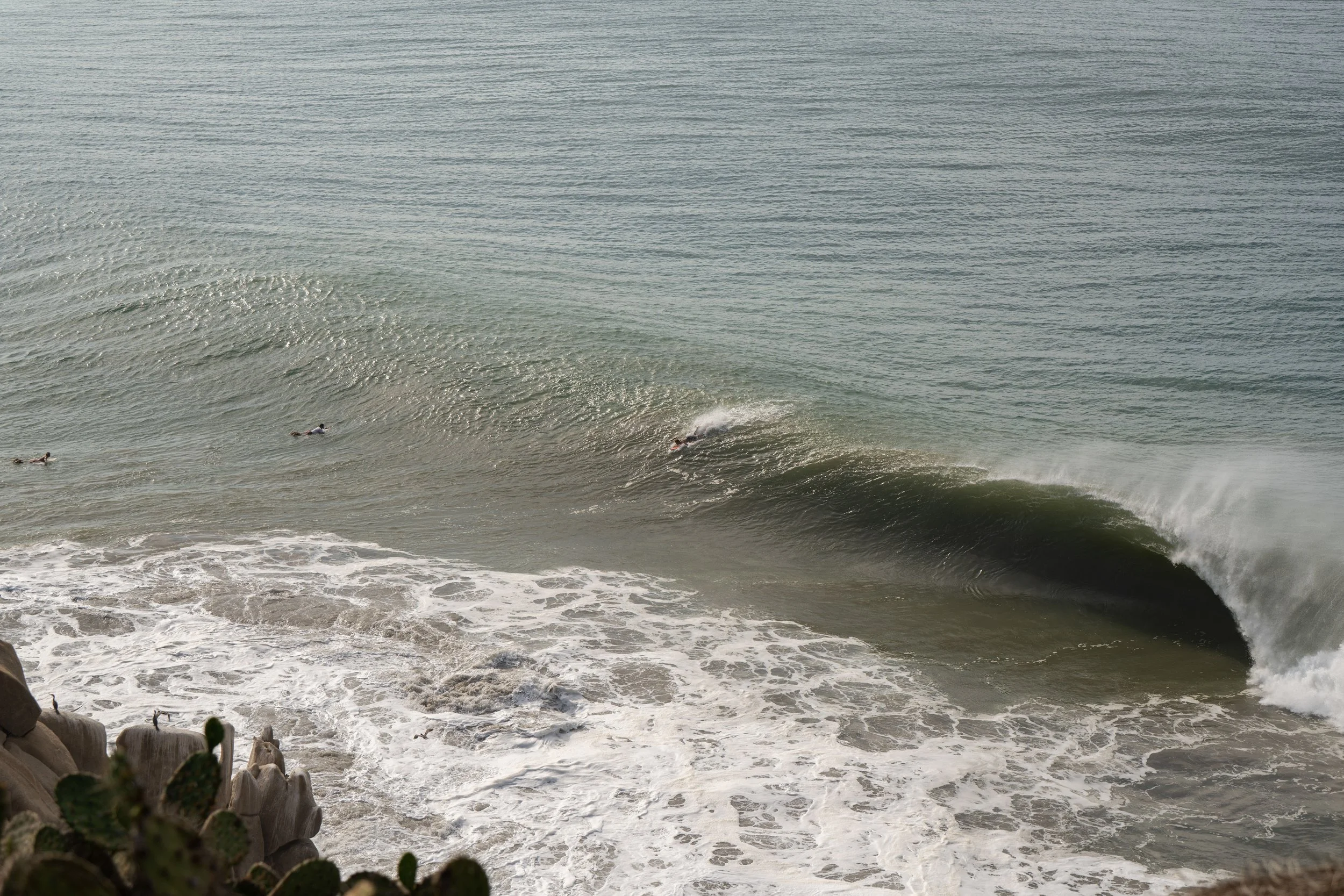 Surfers riding ocean waves near rocks in the foreground.