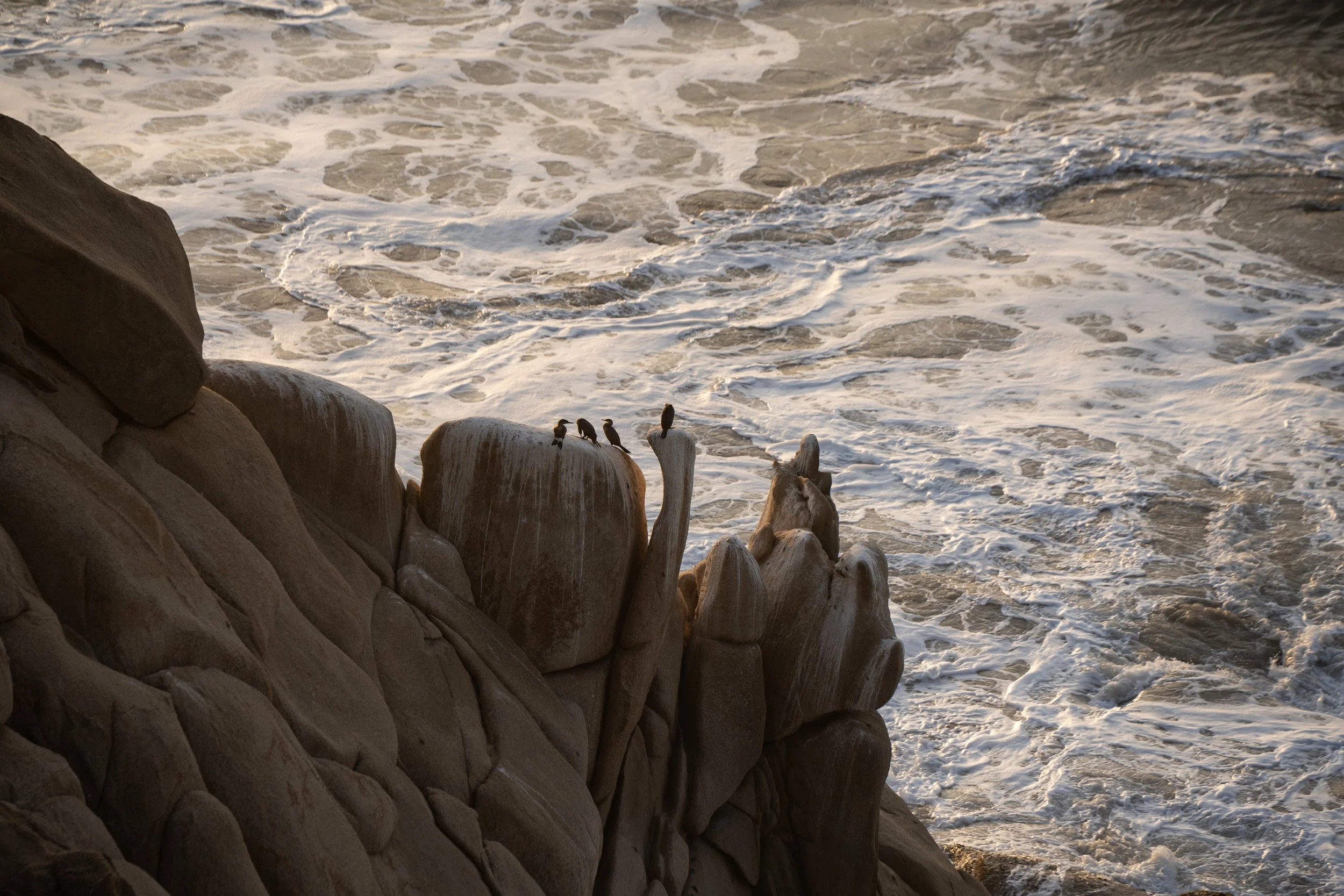 Seaside scene with large rocks near the shore and waves crashing against them, creating foam and spray.
