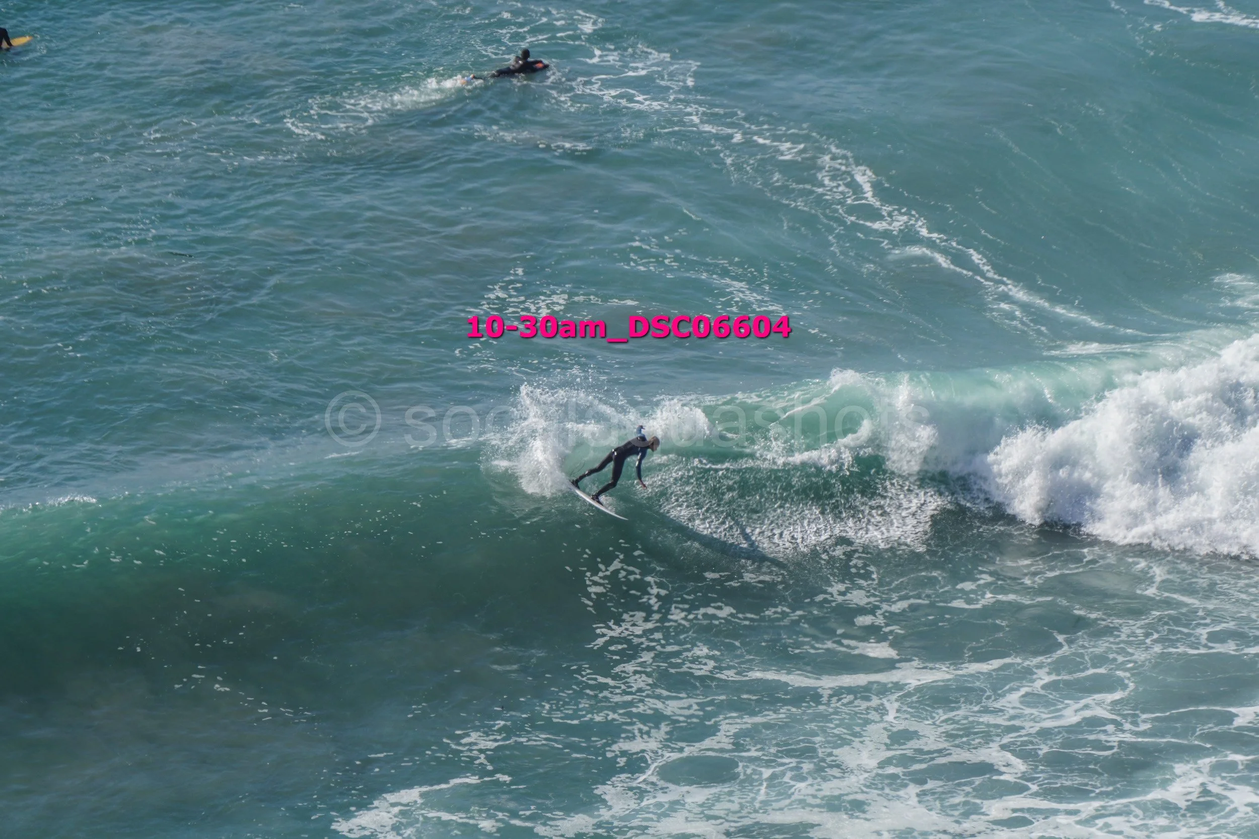 A surfer riding a wave while another surfer paddles in the background in the ocean.