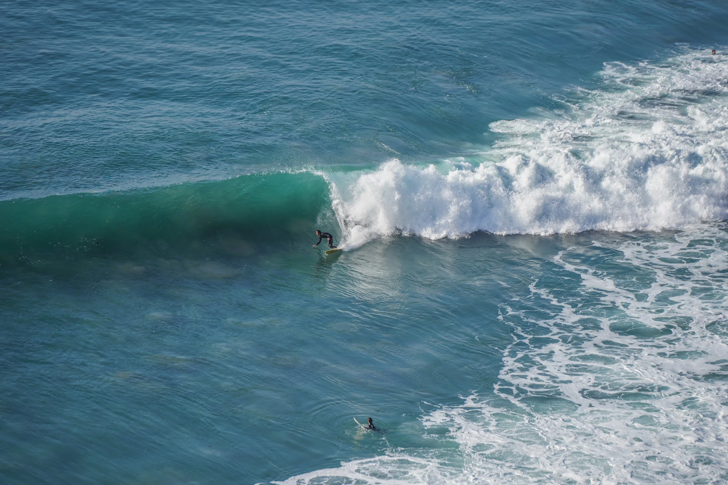 A surfer riding a wave in the ocean, with another surfer swimming in the water below.