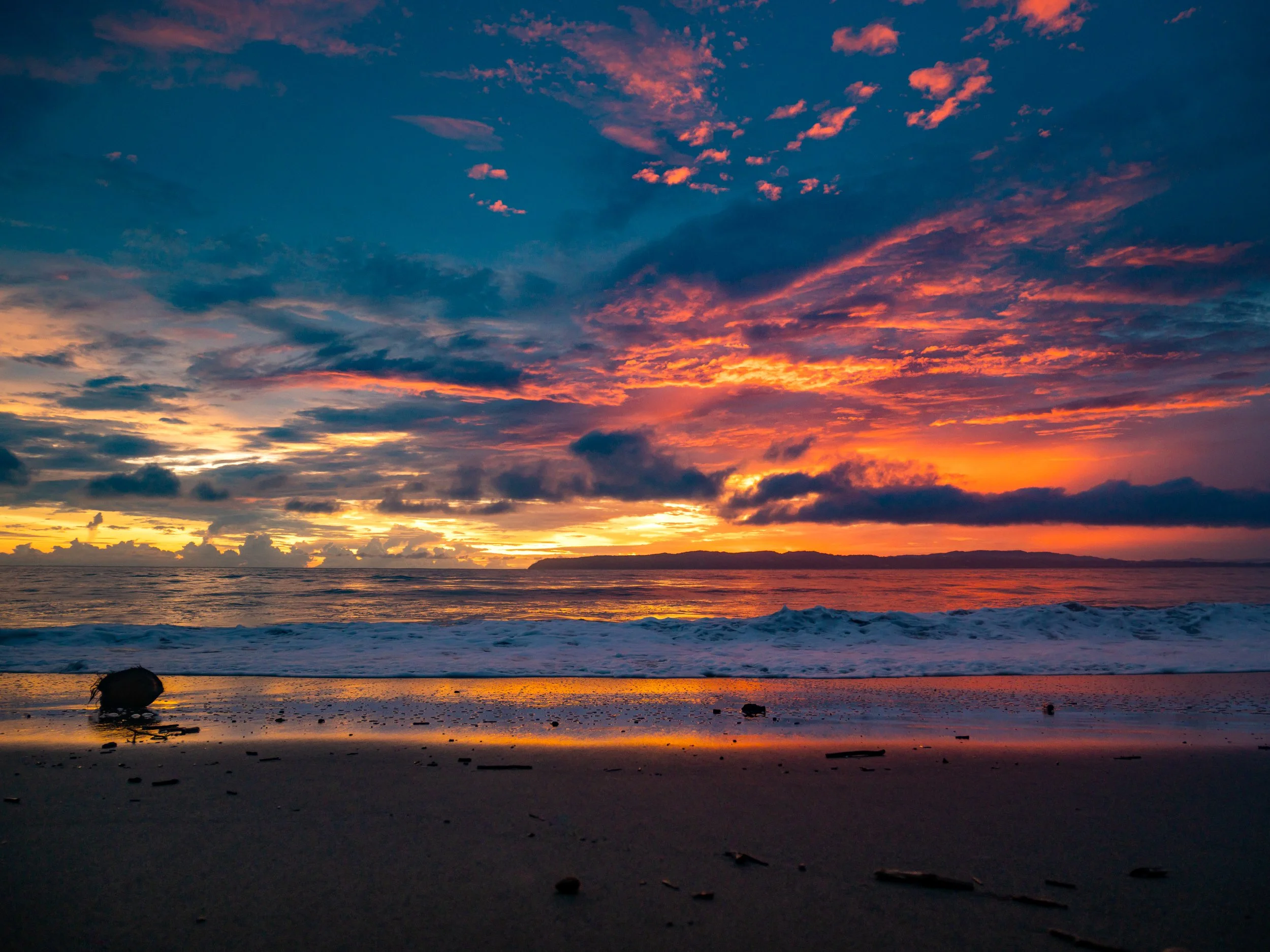 Sunset over the ocean with colorful sky, reflecting on wet sandy beach with debris and small waves.