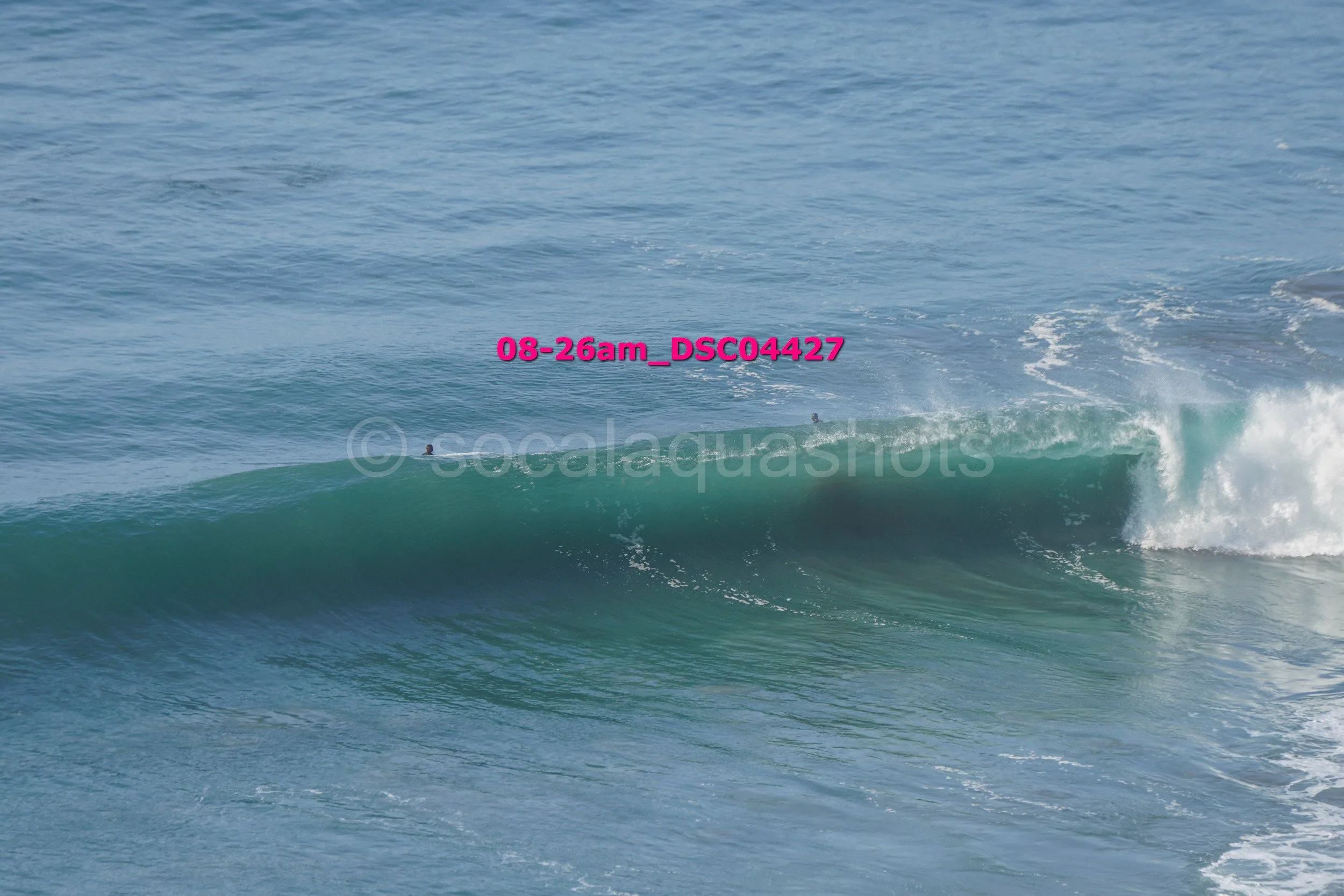Ocean waves with two surfers visible, one on the wave and one behind, under a clear sky.