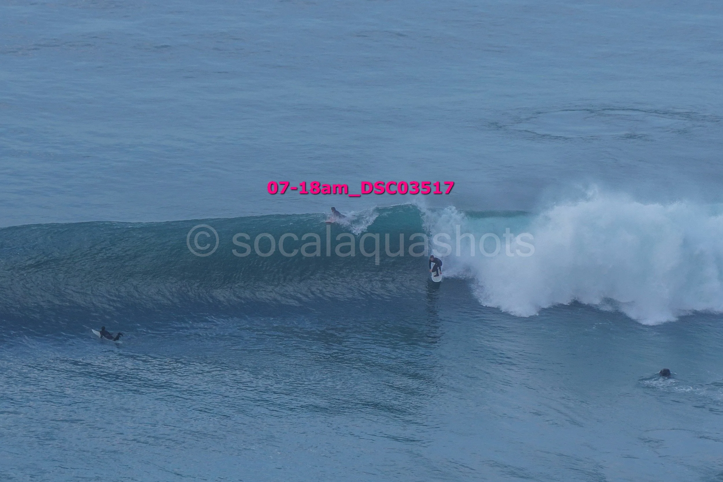 Multiple surfers in the ocean riding and waiting on large waves with visible white crests during daylight, water appears blue under clear skies.