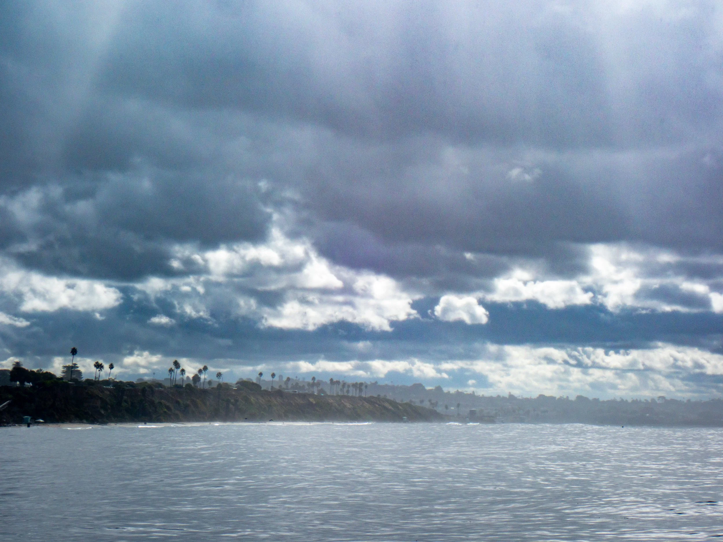 Overcast sky with dark clouds over a coastal landscape with palm trees, shoreline, and calm water.