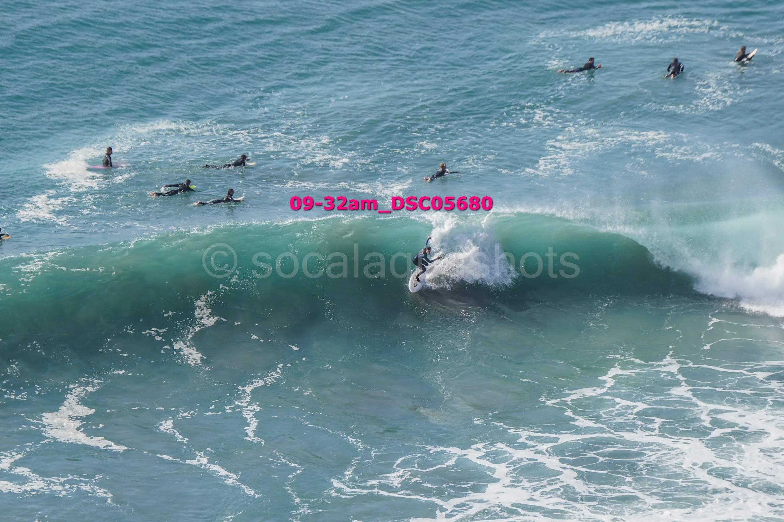 Surfer riding a large wave with multiple surfers in the water nearby.