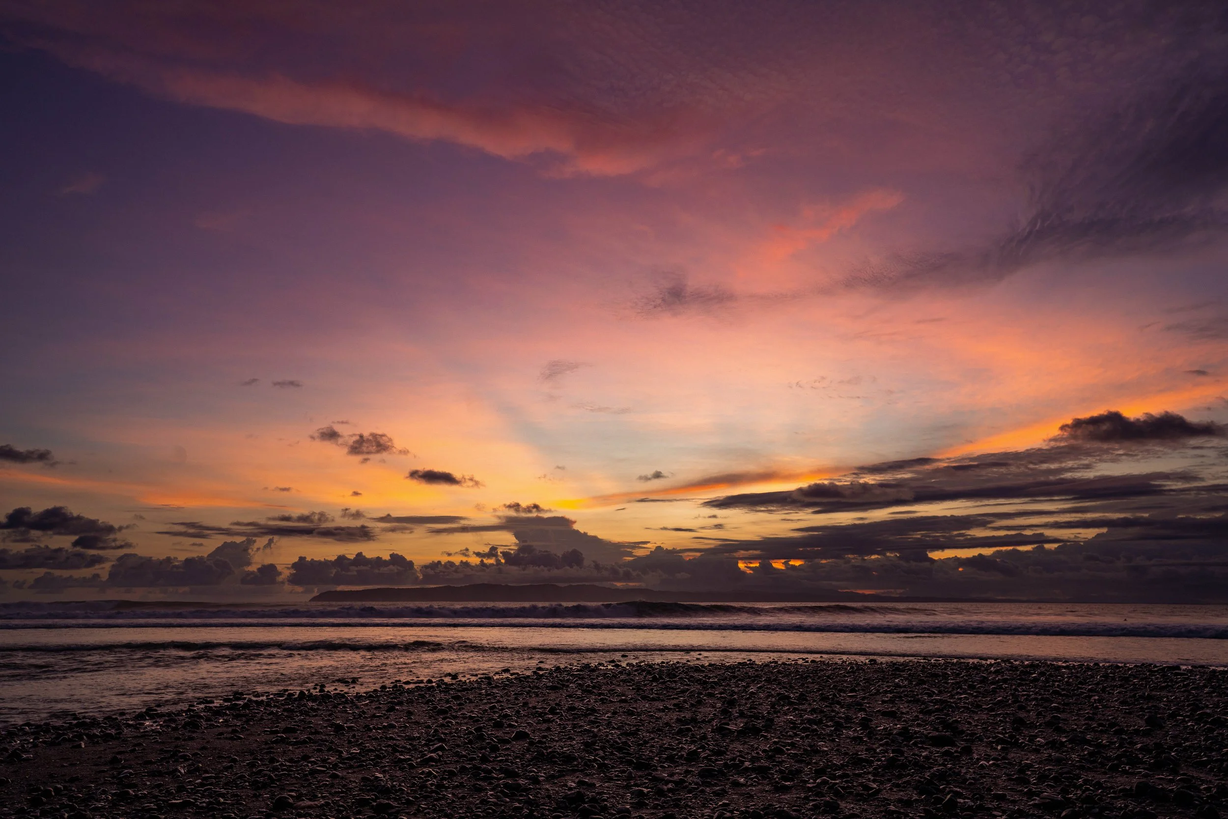 Sunset over the ocean with colorful pink, purple, and orange clouds and a dark rocky beach in the foreground.