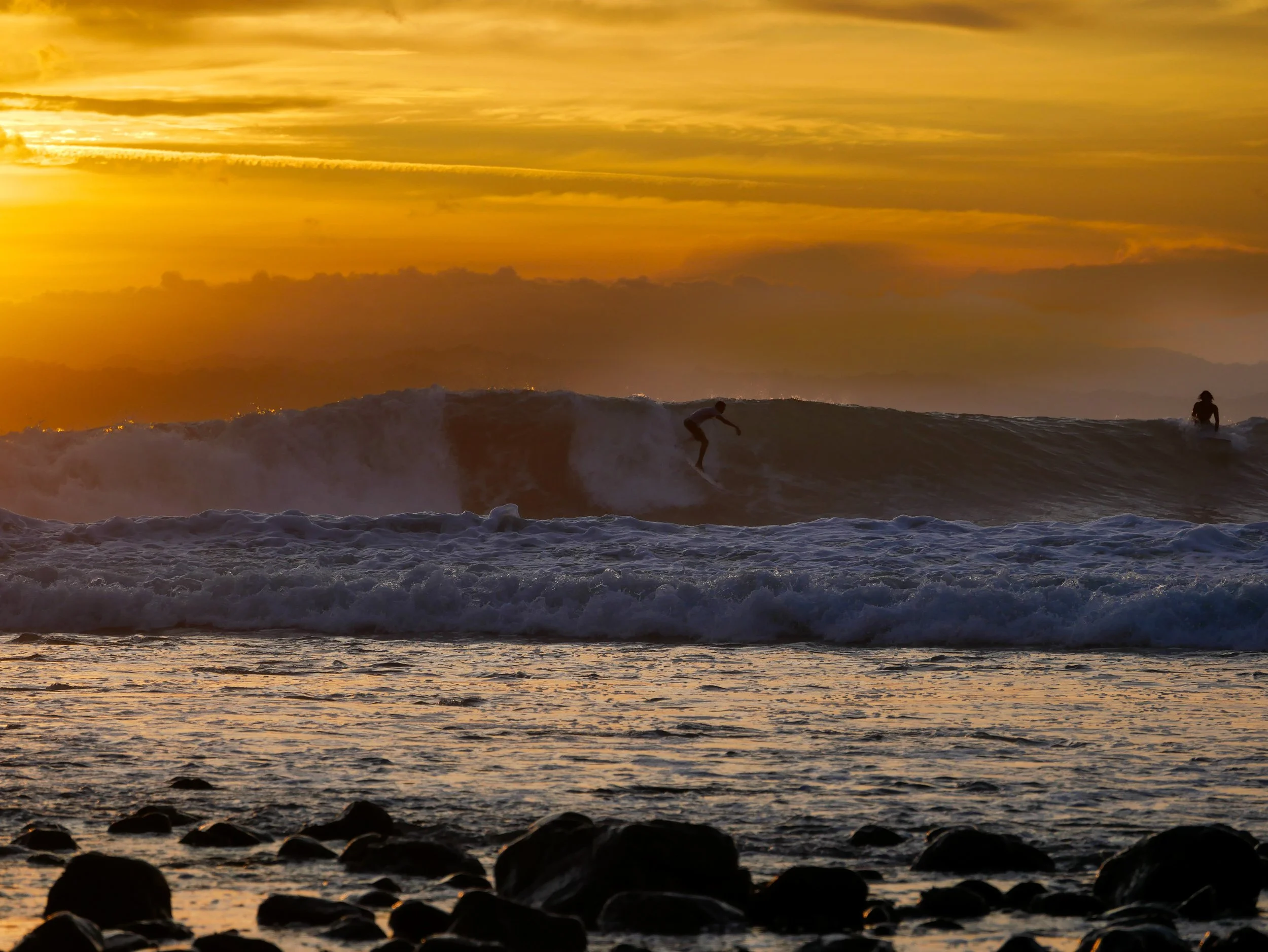 Two surfers riding ocean waves during a golden sunset with rocks in the foreground.
