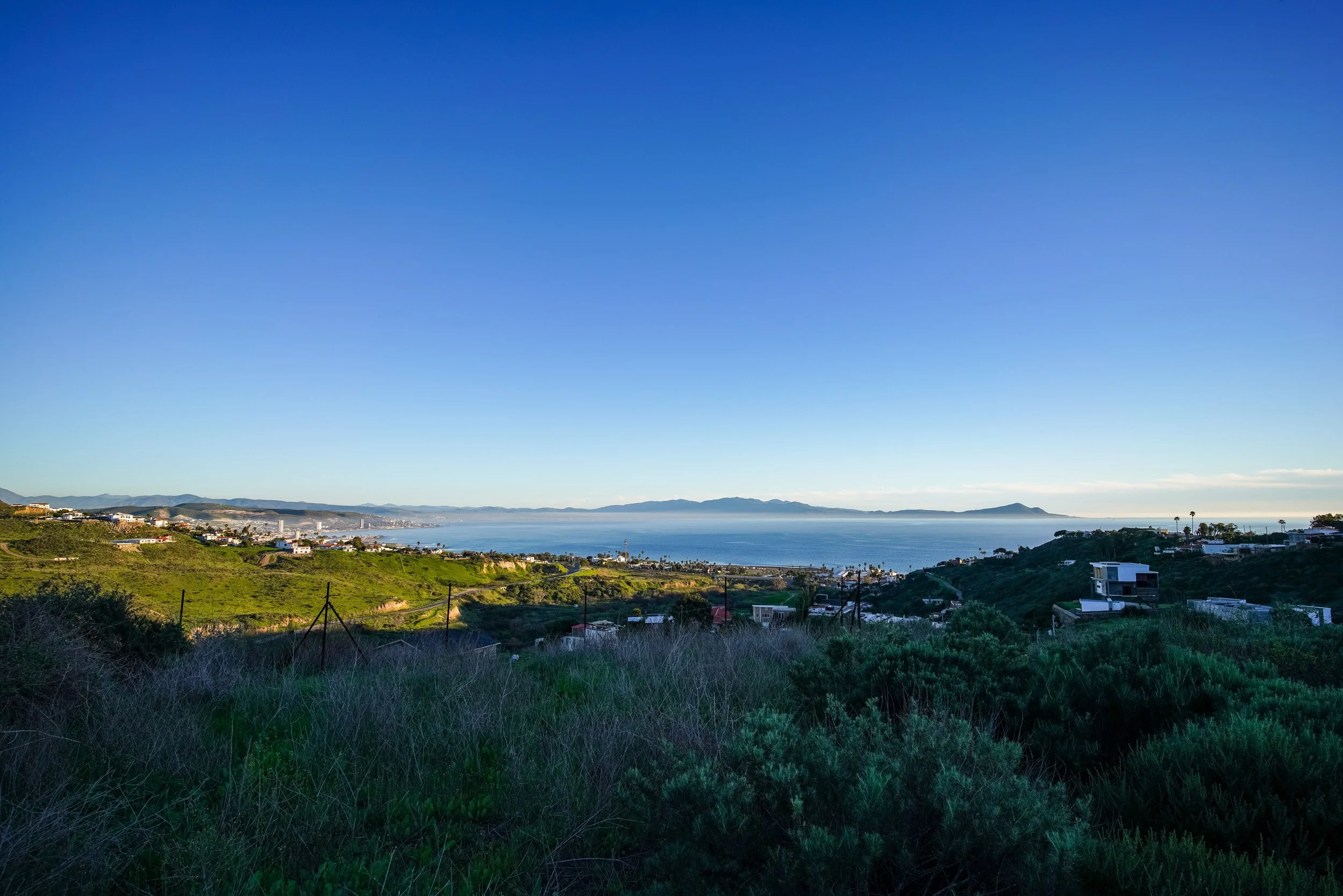 View of a coastal landscape with green hills, houses, and a calm blue ocean under a clear blue sky.