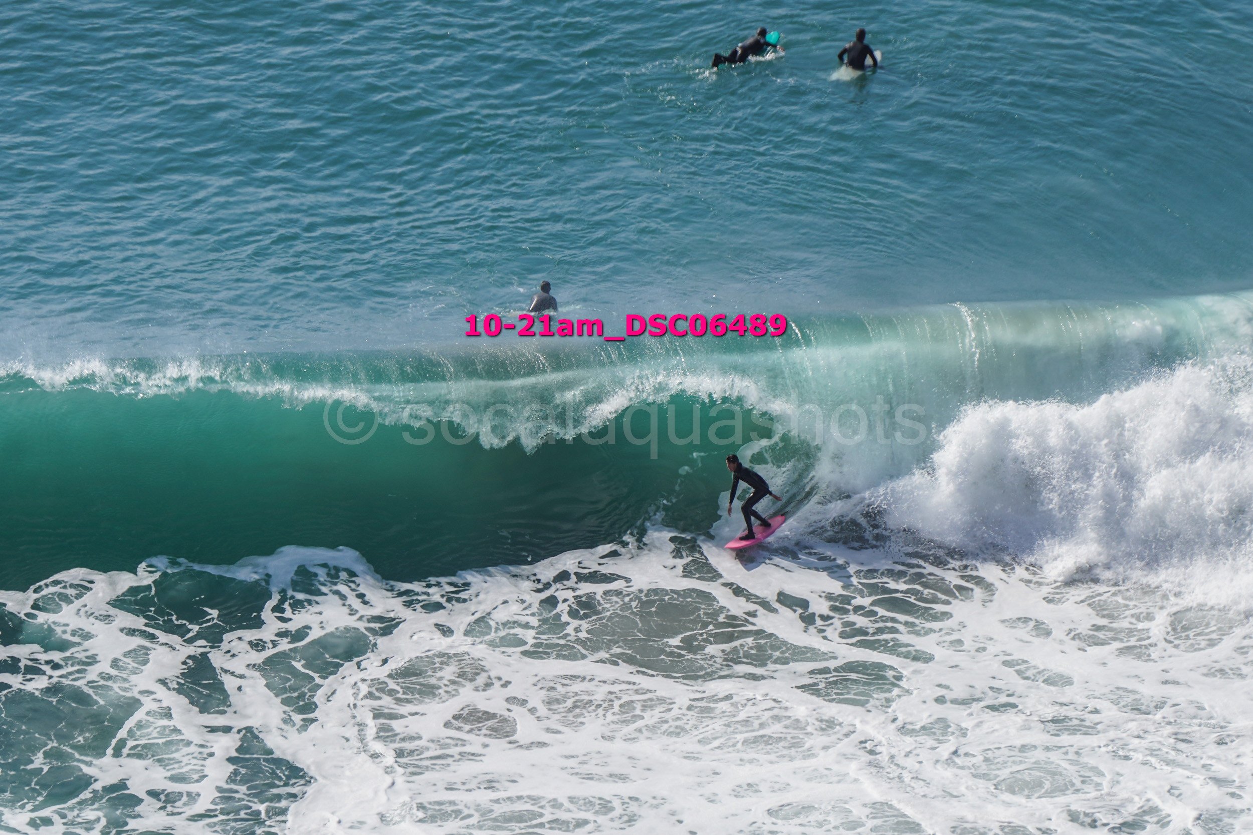 A person surfing on a pink surfboard riding a wave, with three other surfers in the distance in the water.