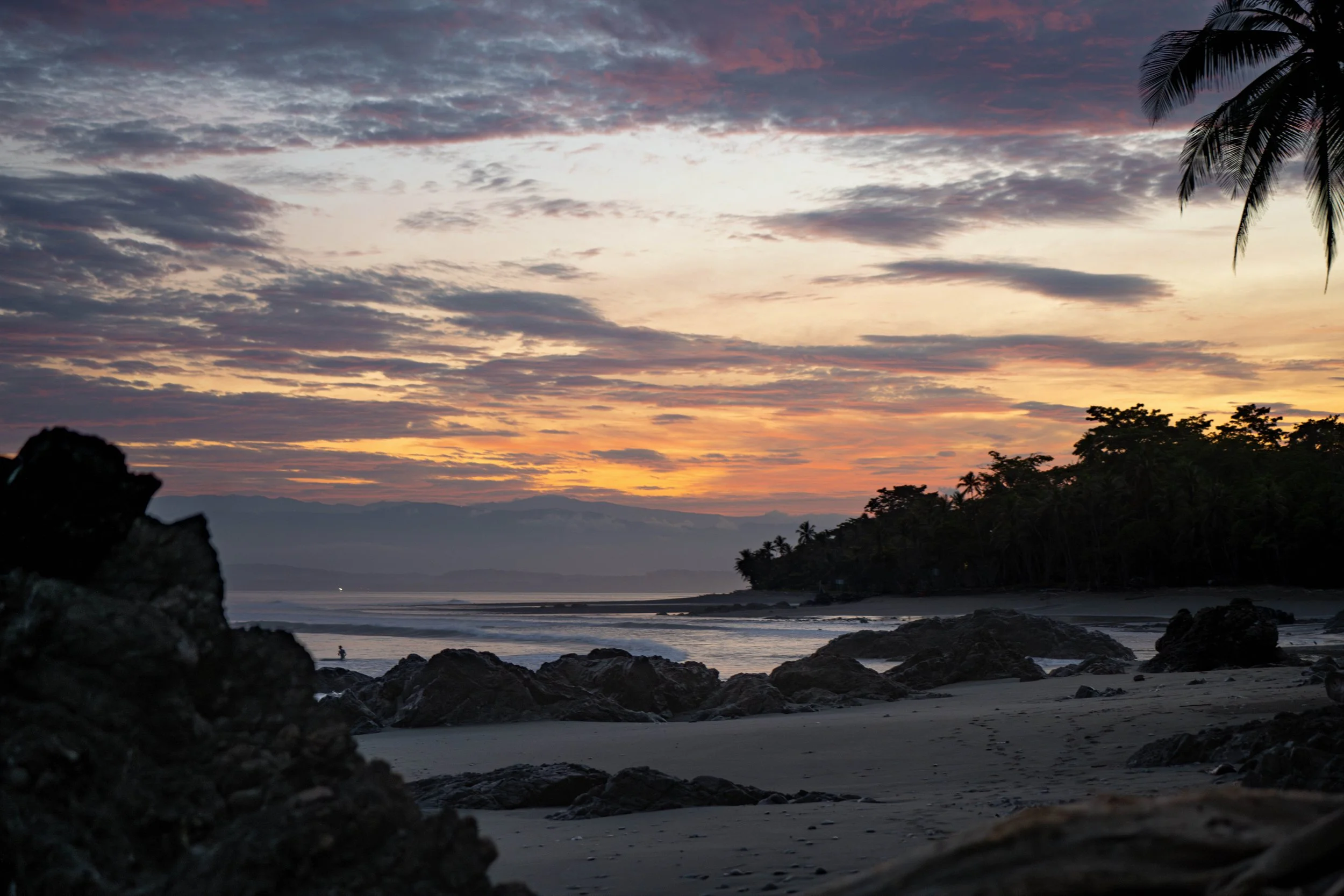 A scenic beach at sunset with colorful clouds in the sky, silhouettes of trees and rocks along the shoreline, and a calm ocean.