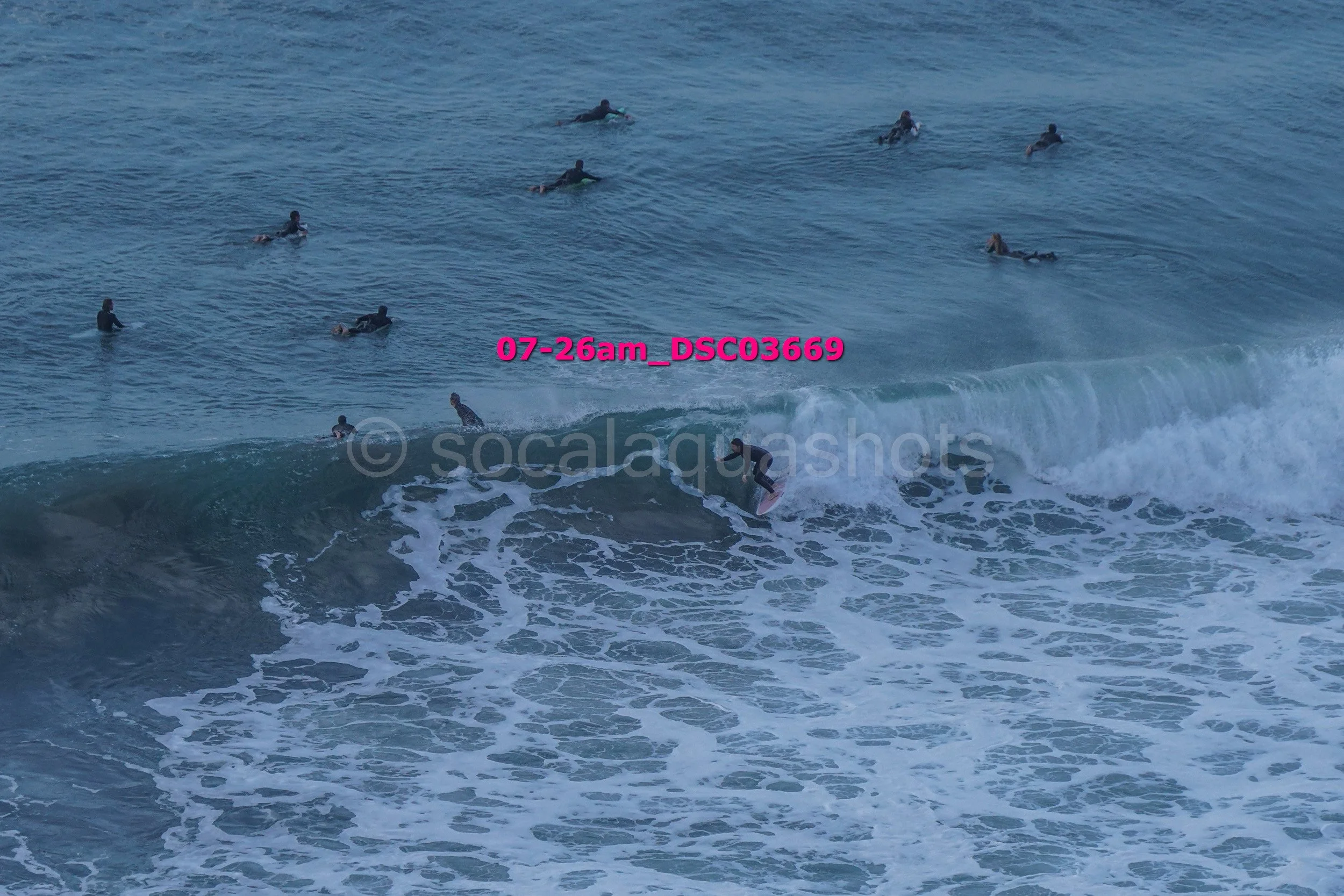 A group of surfers in the ocean, some riding waves and others waiting in the water, with one surfer on a wave in the foreground.