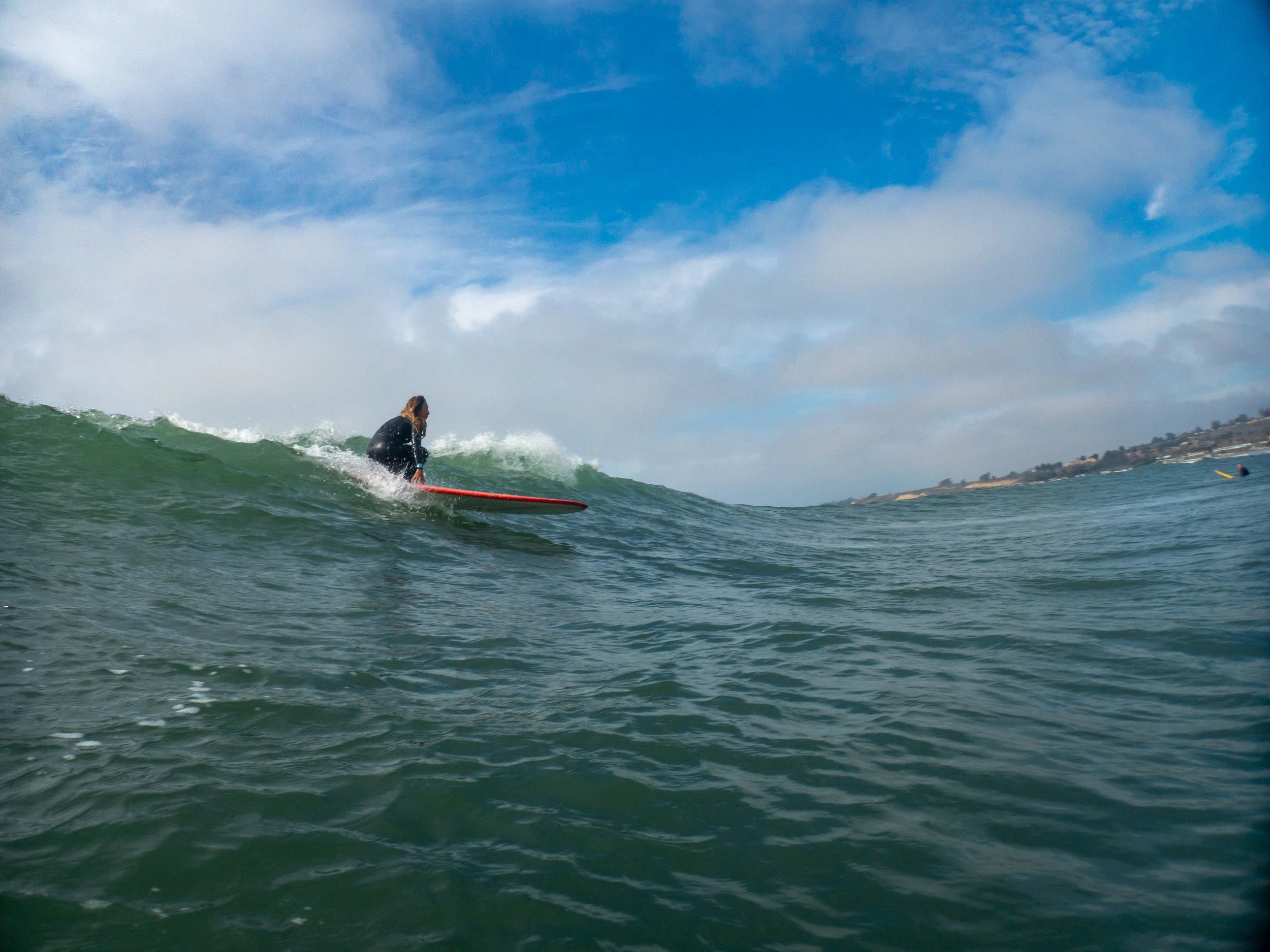 A person surfing on a wave in the ocean, with a partly cloudy sky and coastal land in the background.