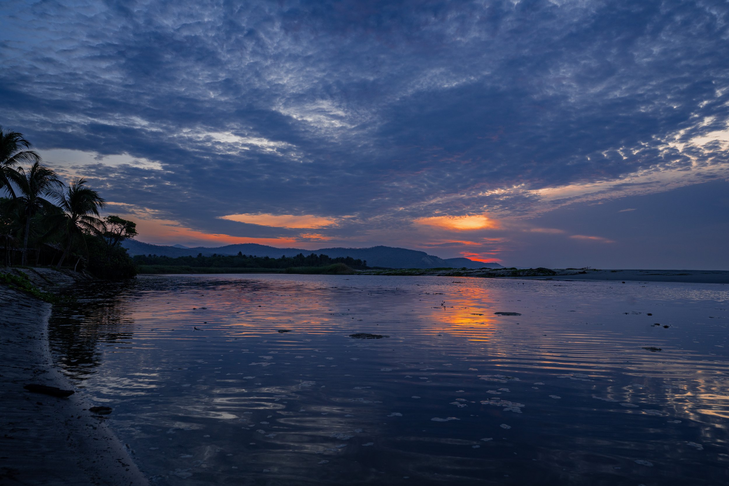 A peaceful sunset over a river with mountains in the background and trees on the left, reflecting the colorful sky.