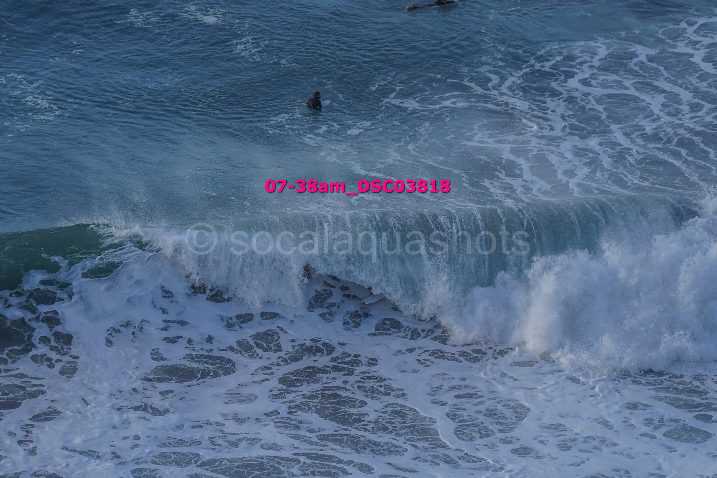 Surfer in a black wetsuit riding a large wave in the ocean with white foamy surf, viewed from above.