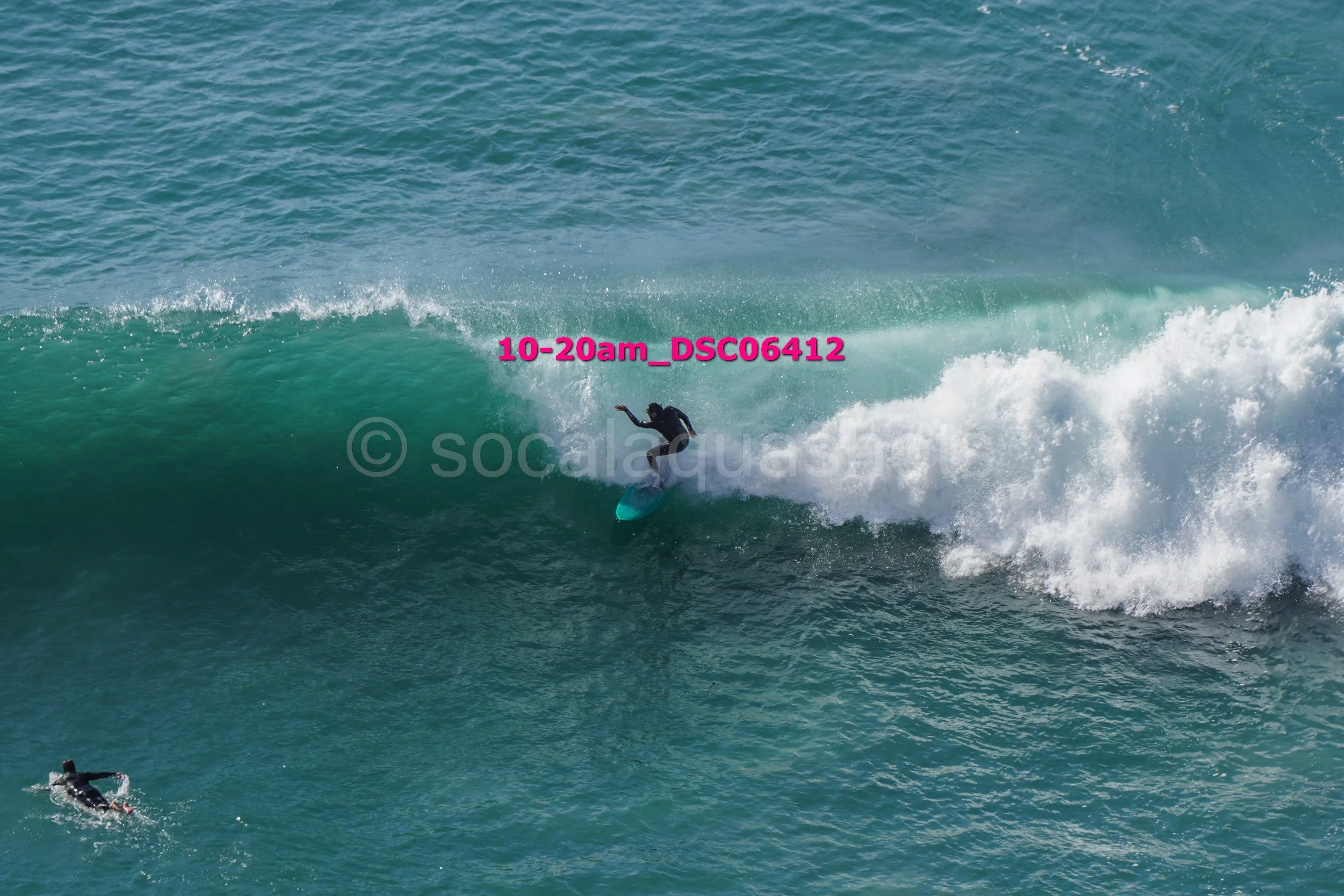 A person surfing on a large ocean wave with another surfer swimming nearby.