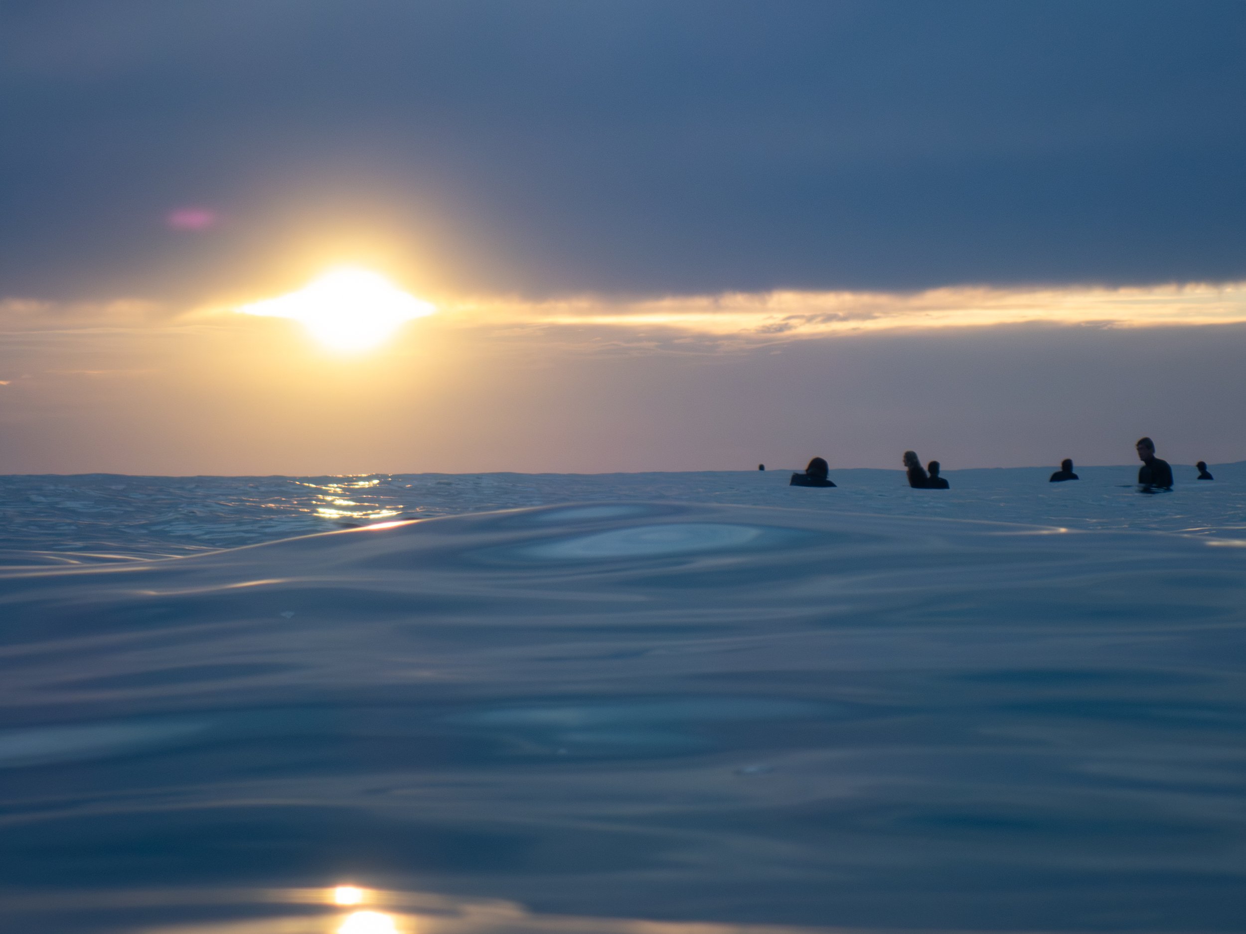People swimming in the ocean at sunset, with dark clouds overhead and the sun low on the horizon.