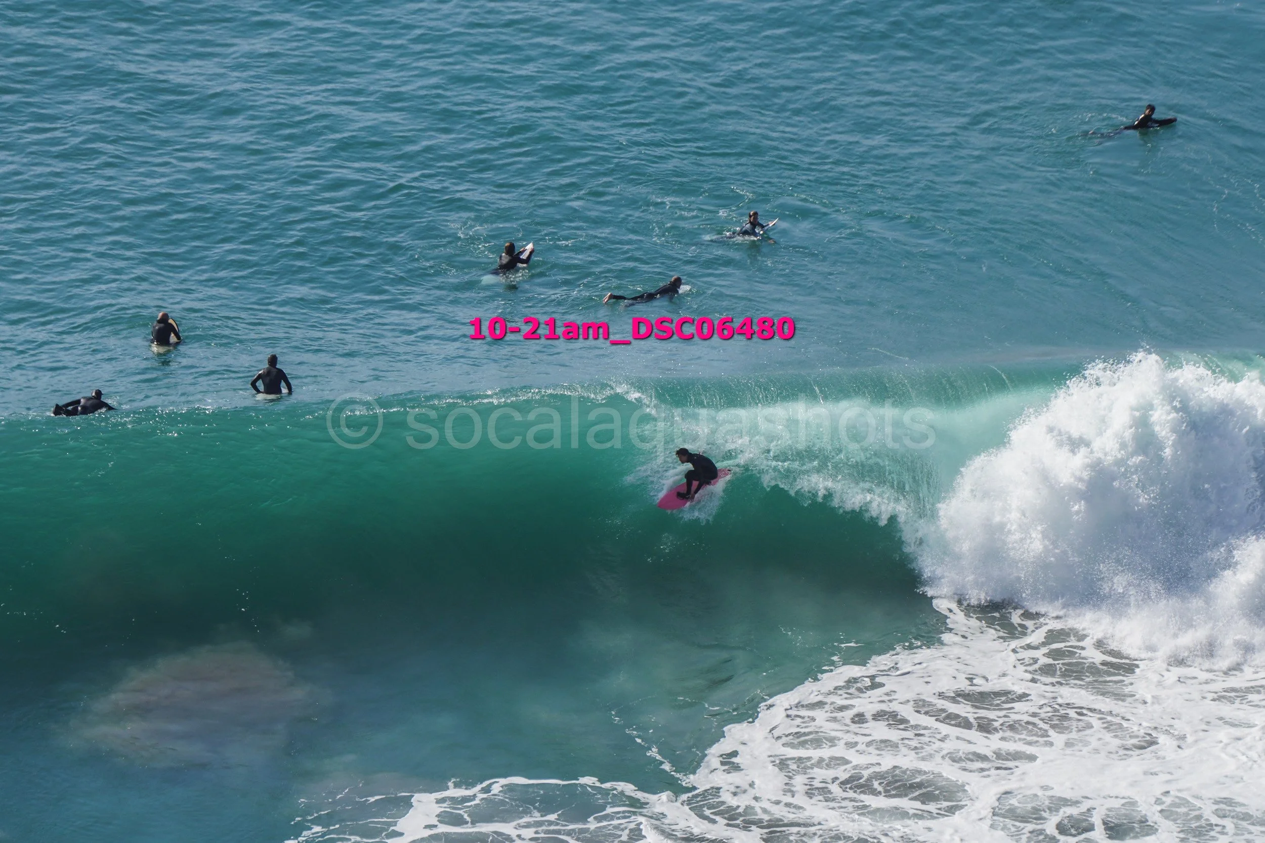 Surfer riding a wave near the coast with multiple people in wetsuits in the water, watching the surf.