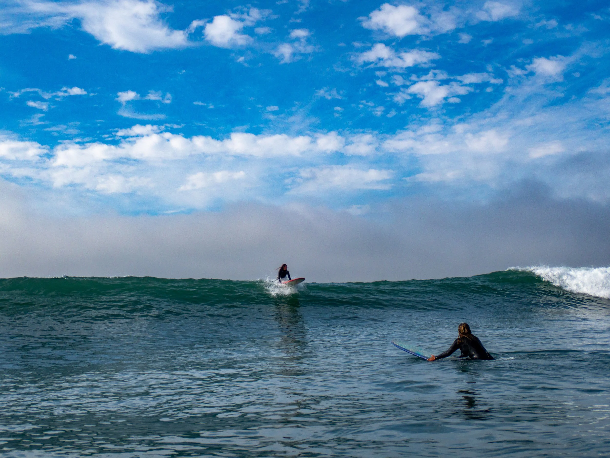 A surfer riding a wave and another person holding a surfboard in the water under a partly cloudy sky.