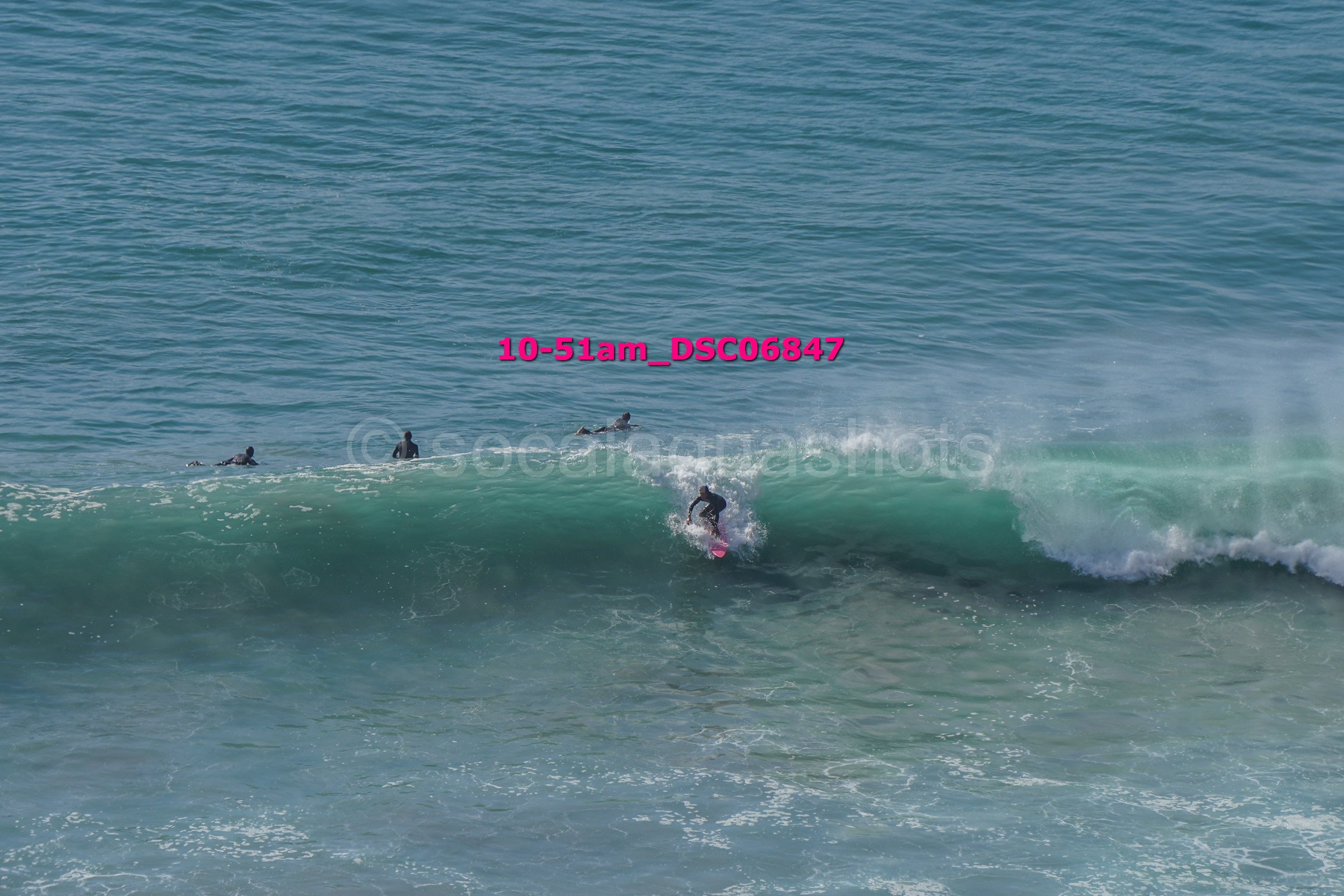 A person surfing on a wave with three other people in the water nearby, on a sunny day at the beach.