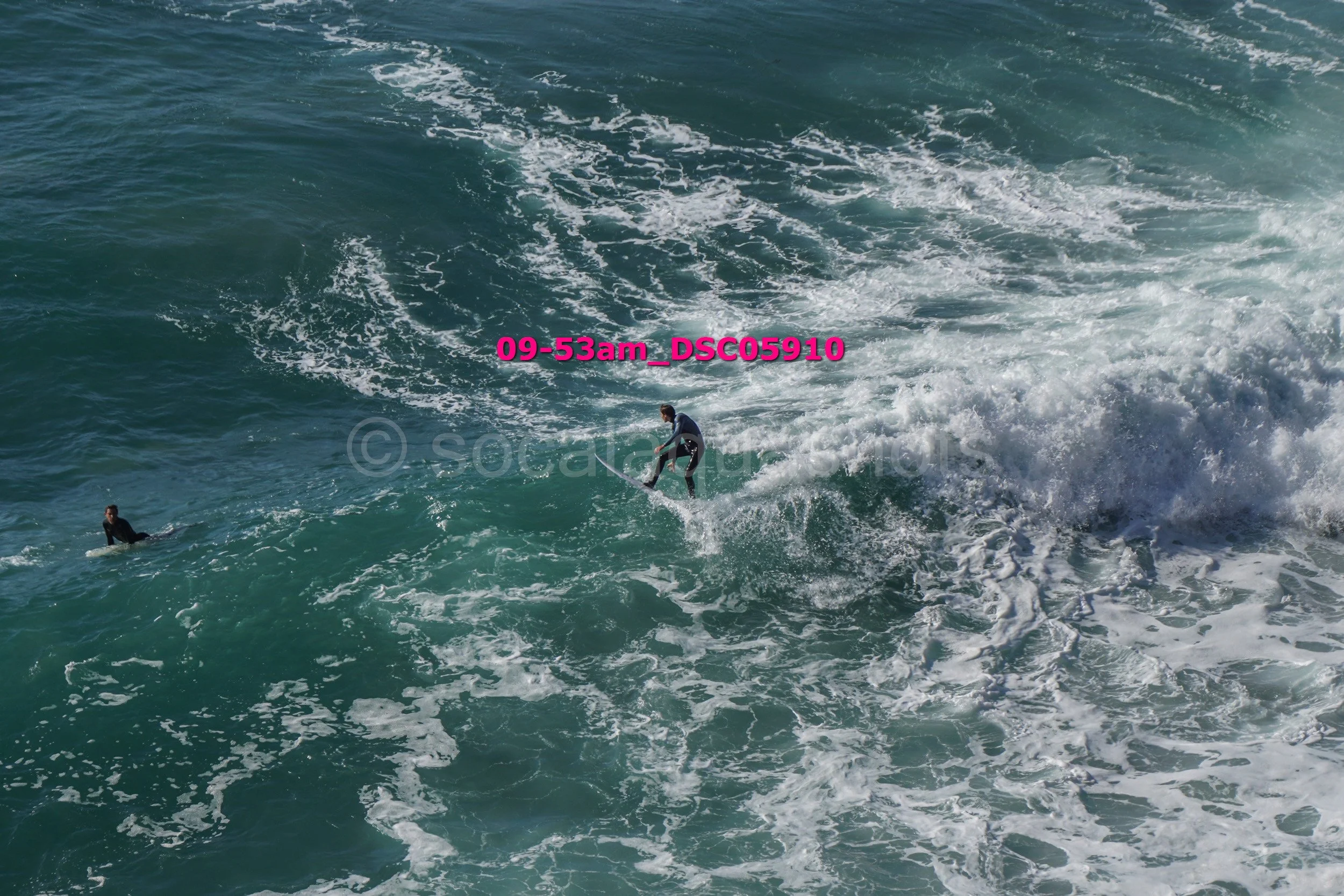 A person surfing on a large ocean wave with another person nearby in the water.
