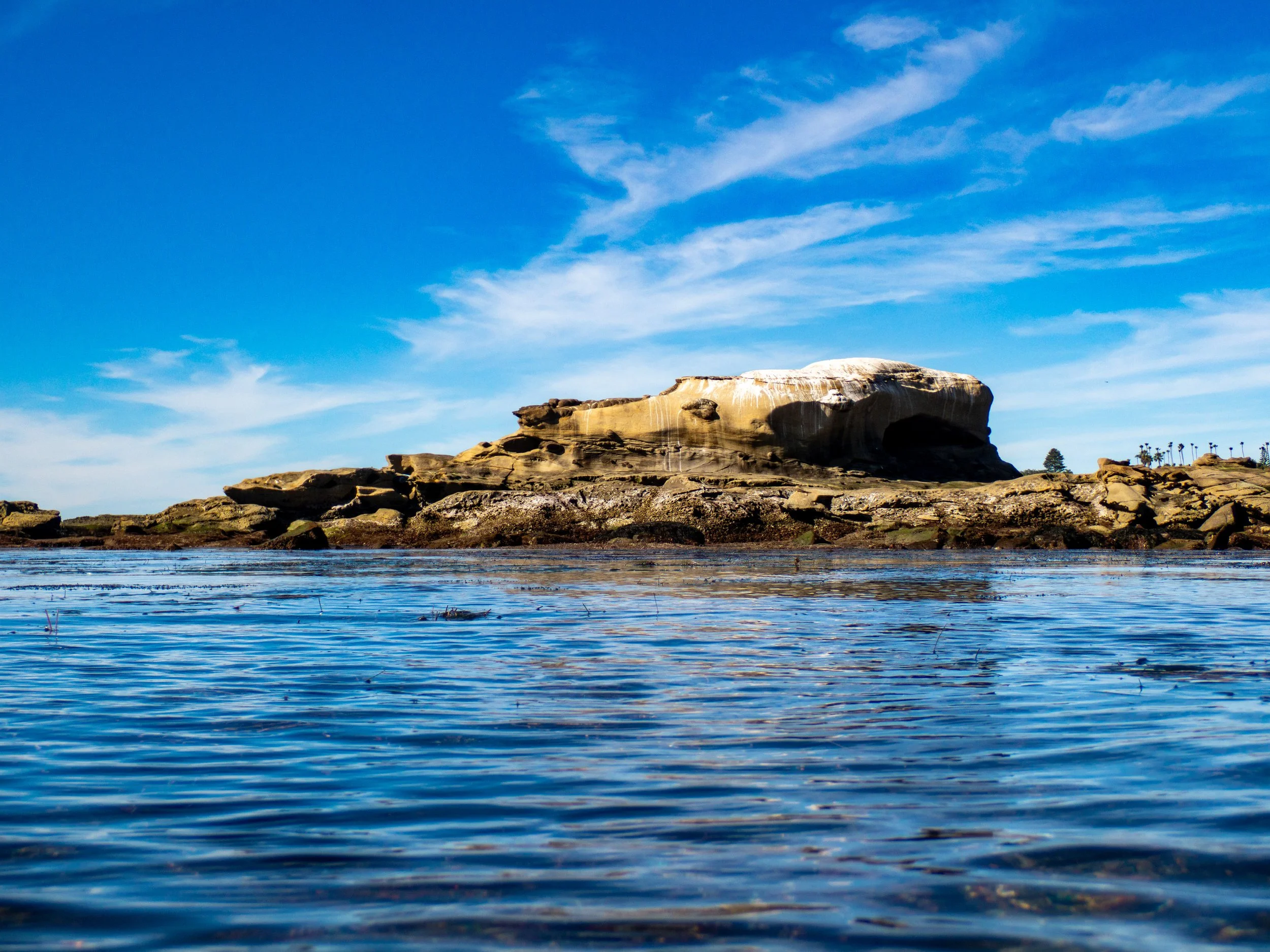 A large rock formation on a coastline with a few trees in the background, under a partly cloudy blue sky, reflected in the water.