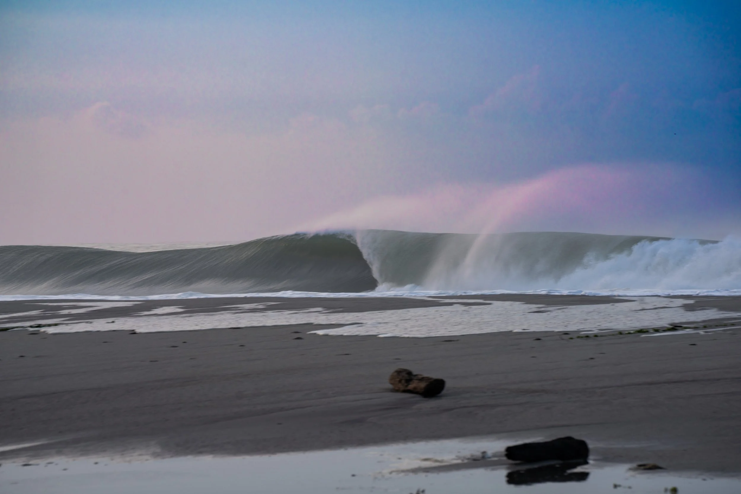 Oceanside beach with large crashing wave, wet sand in foreground, cloudy sky with a hint of rainbow.