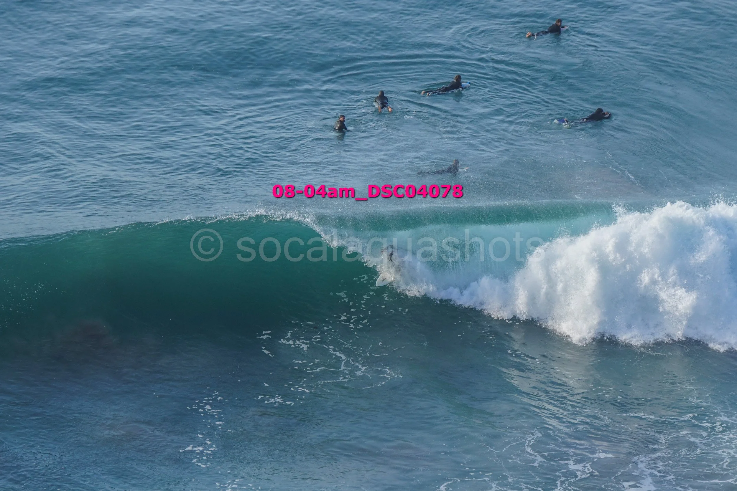 Group of surfers in the water, with one riding a wave and the others swimming or waiting nearby.