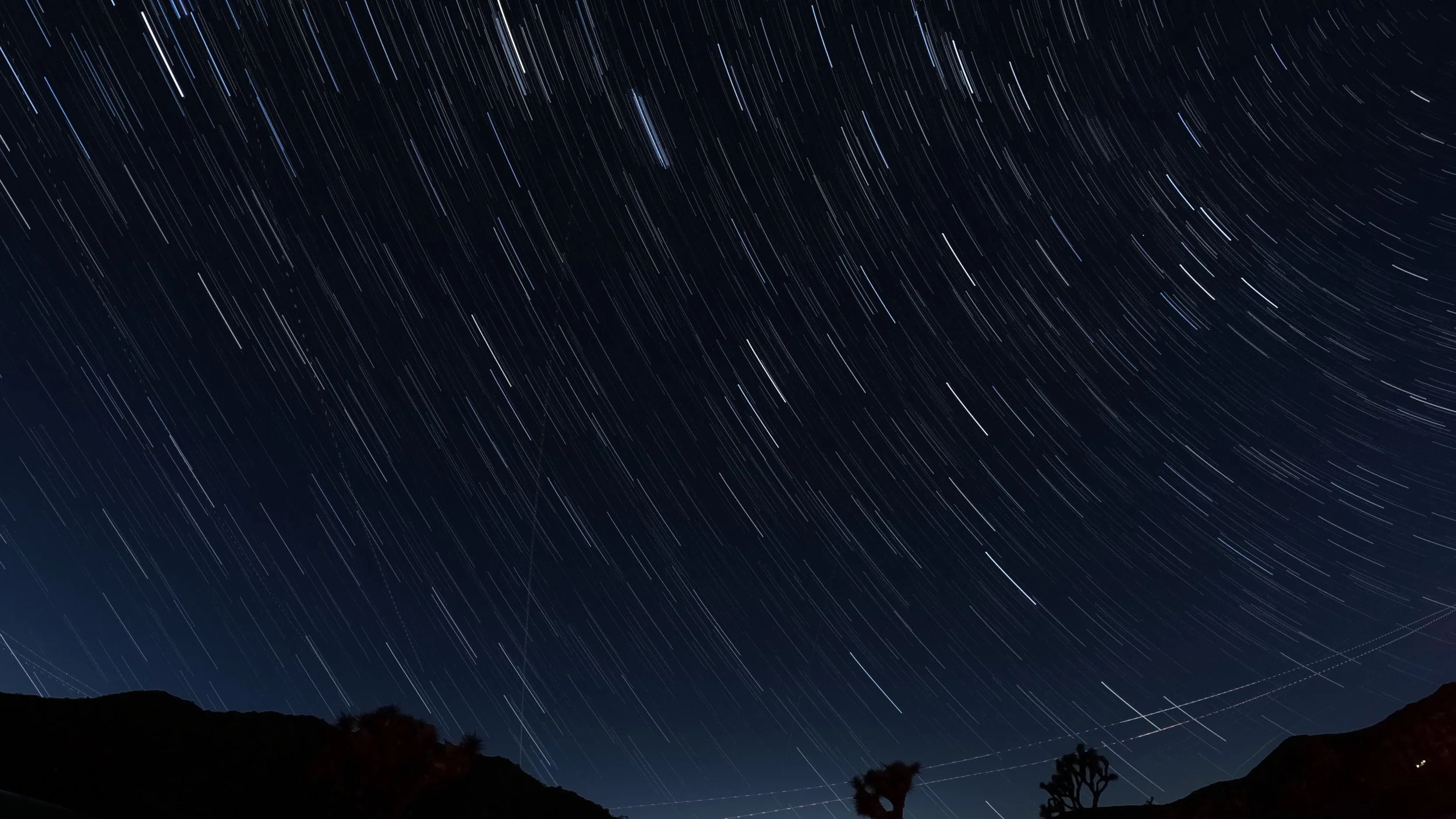 Star trails streak across the night sky over a dark mountain landscape with silhouetted trees.