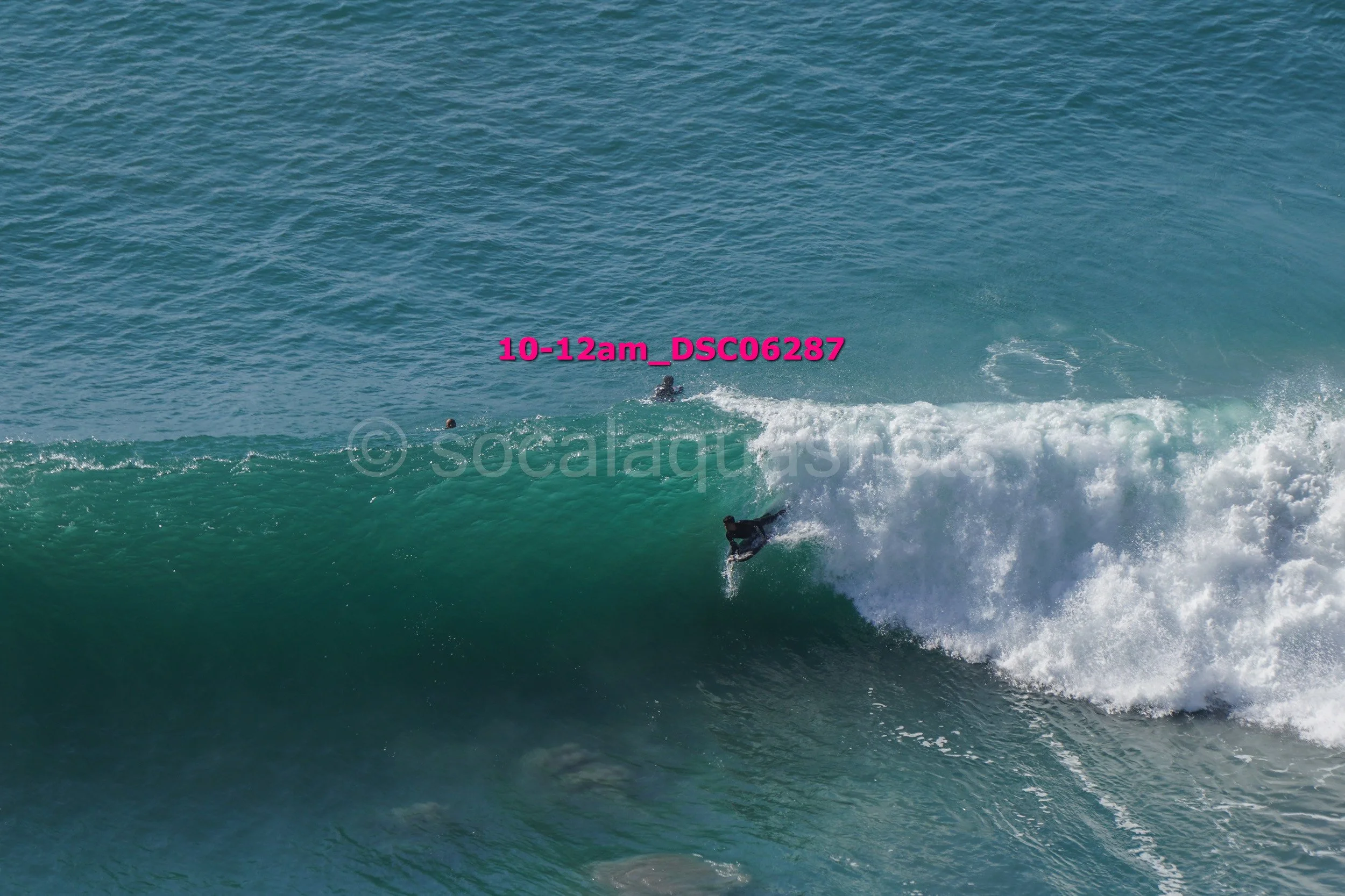 A person surfing on a large ocean wave with an offshore water background, wearing a wetsuit, with two other individuals visible in the water nearby.