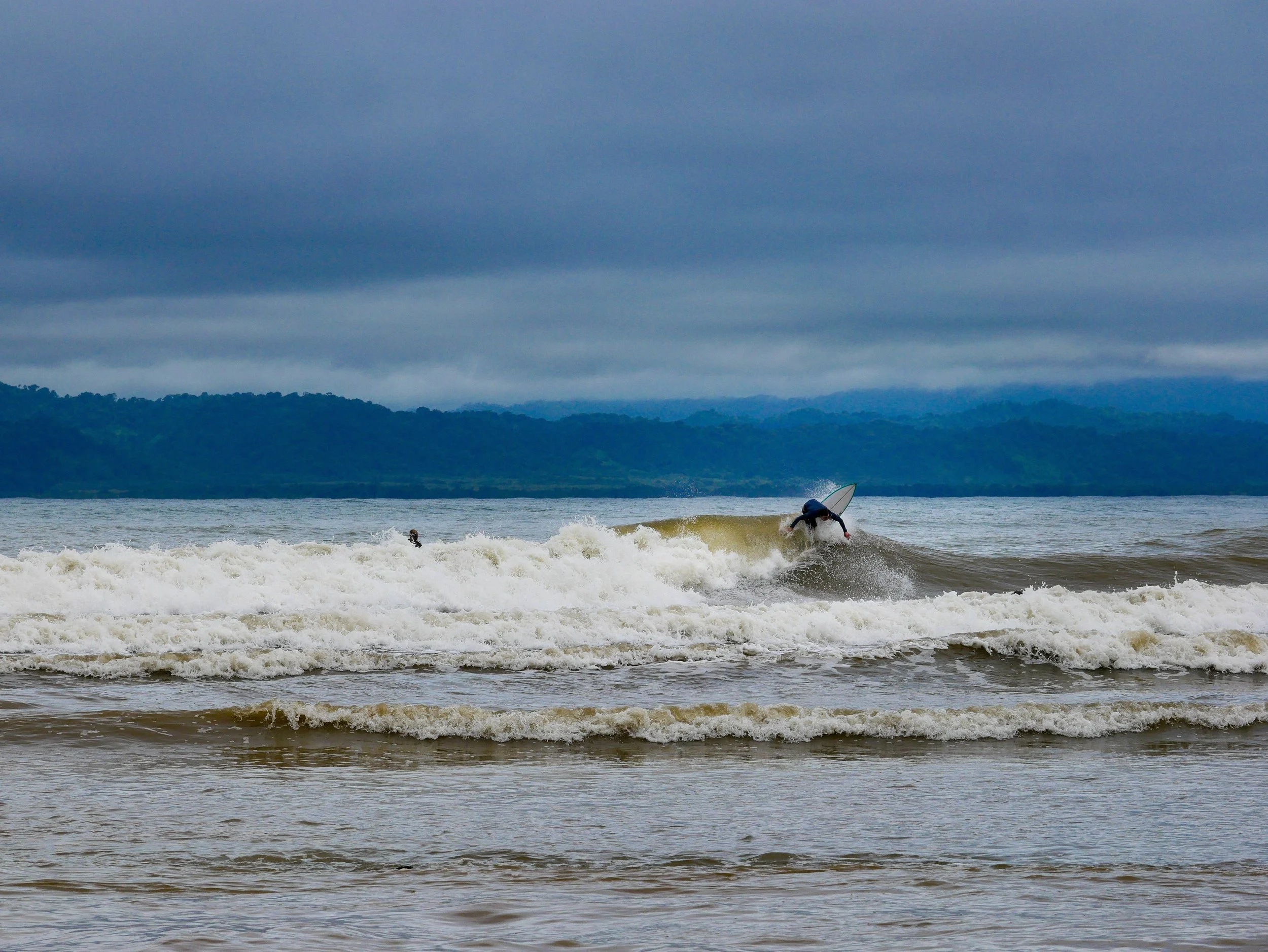 Surfer riding wave in ocean with cloudy sky and distant forested hills.