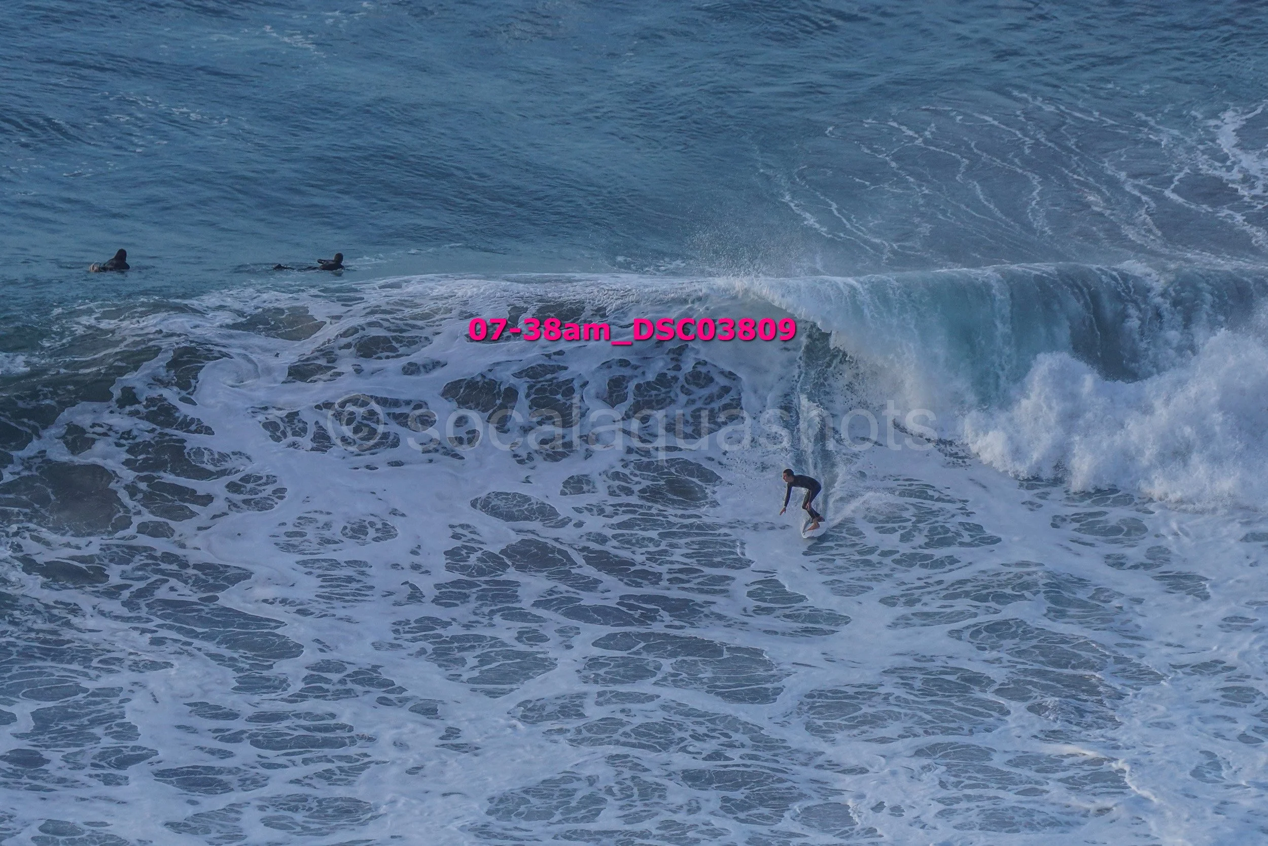 A person surfing a large wave in the ocean with three other surfers visible in the water nearby.