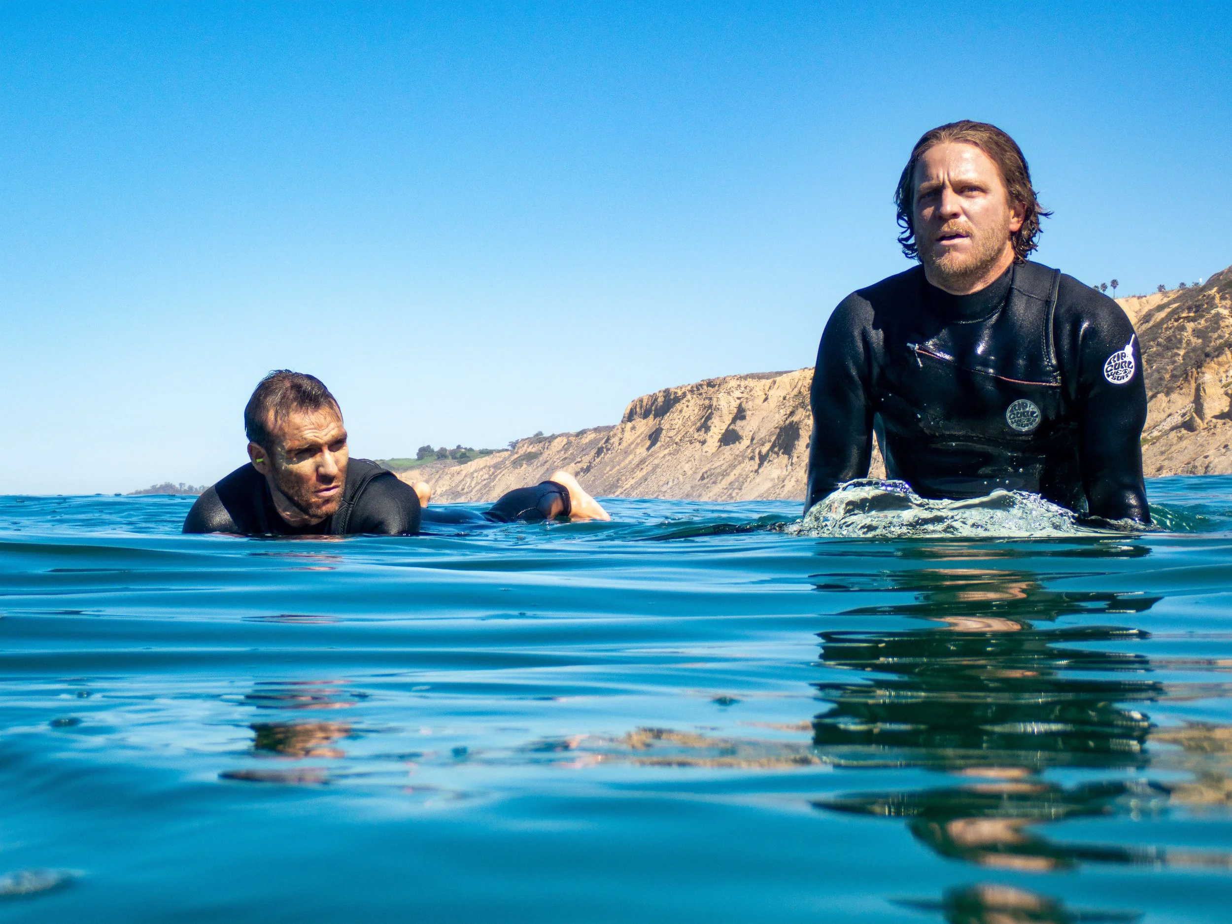 Two men in wetsuits are in the ocean, with one man on the right looking towards the camera and the other on the left swimming beneath the surface. There are cliffs and clear blue sky in the background.